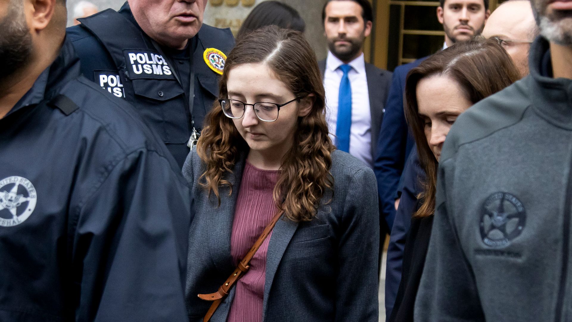 A young woman with her head down, large glasses, wearing a coat and purse, surrounded by people, some in suits, some in police guard uniforms. 