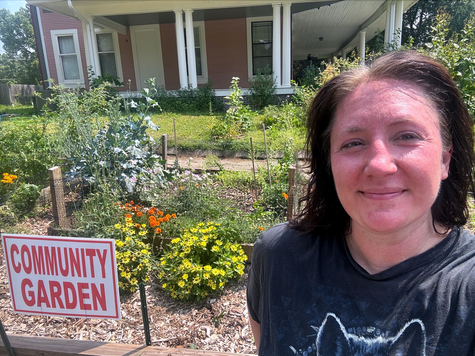 Smiling woman in a dark shirt takes a selfie in front of a pink house with white columns, beside a community garden with yellow and orange flowers and a sign.