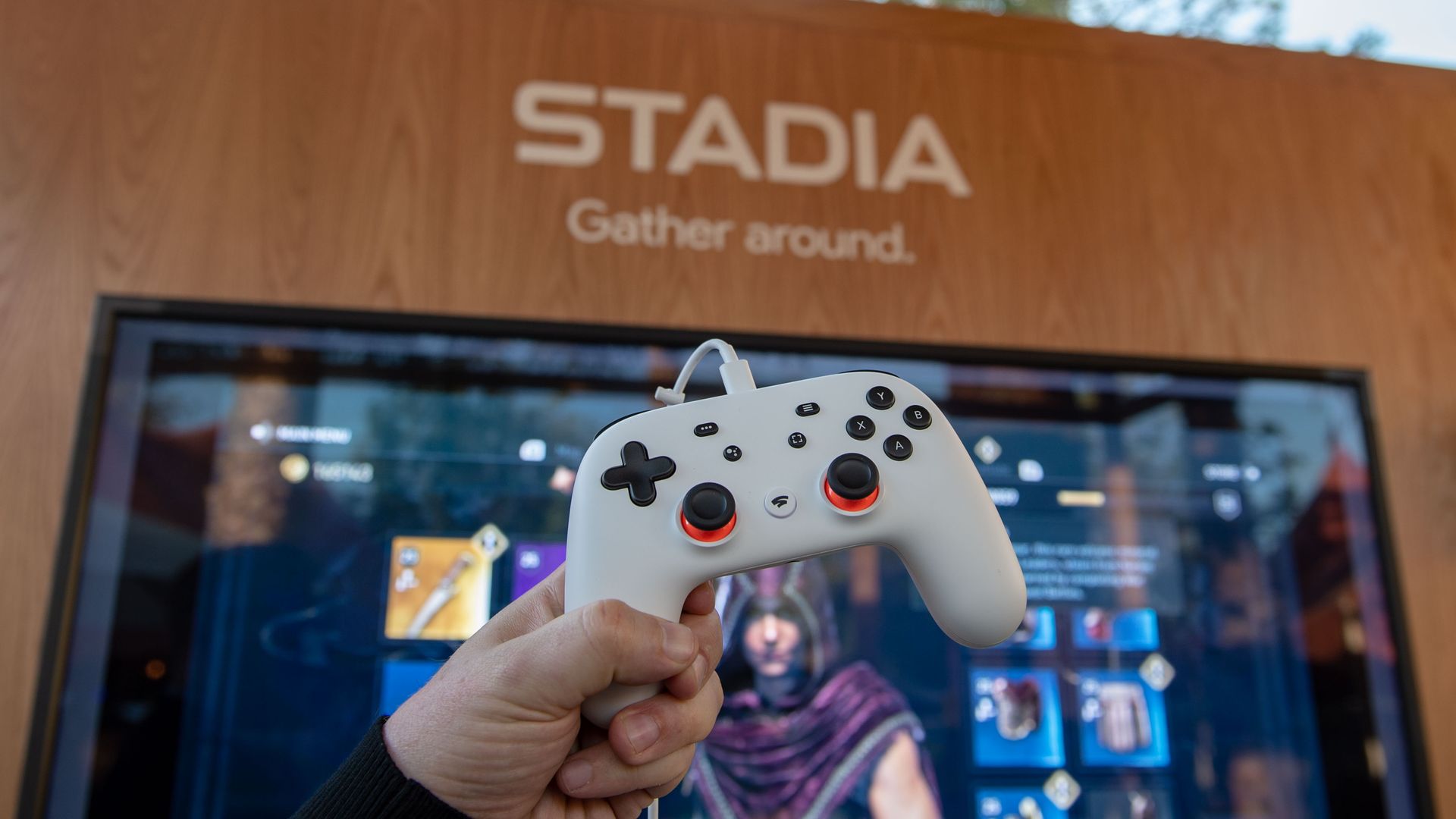 Photo of a person's hand holding a white game controller. A kiosk with the word Stadia is in the background.