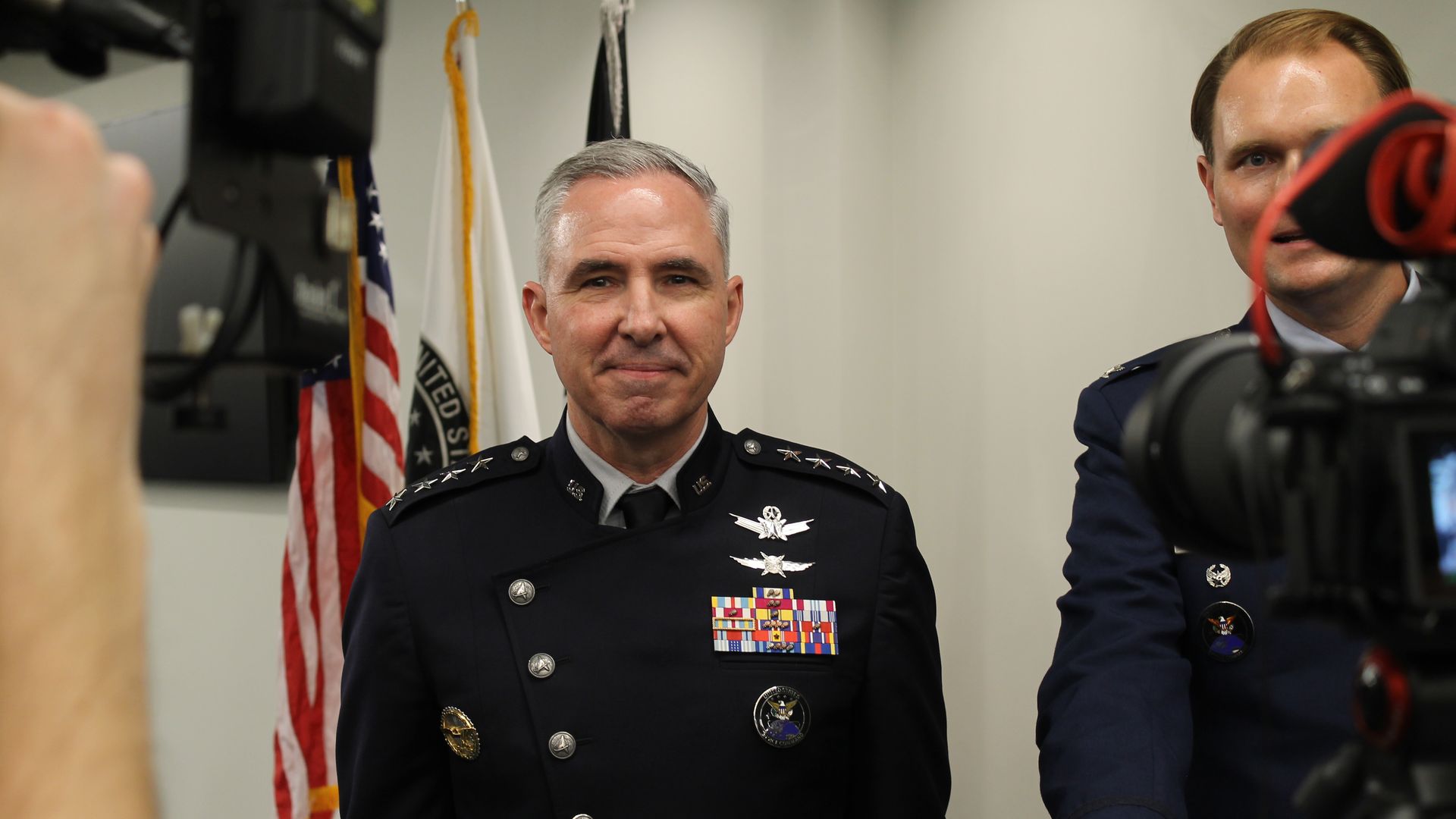 Center: a senior police officer in dress uniform with ribbons and badges, standing before flags. A microphone blocks part of another officer on the right; photographer's hand in foreground.