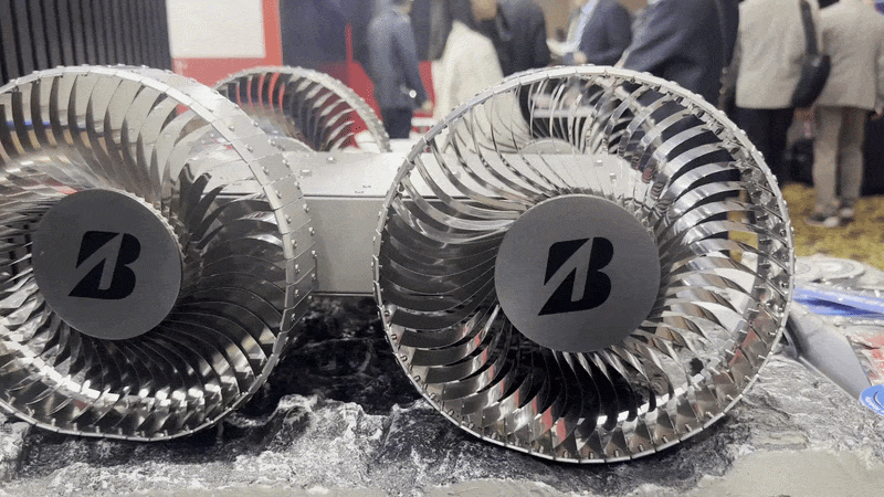 A small metal-gray box, around 10 inches long and four inches wide sits on a display table. The box has four shiny metal springy wheels that look a bit like computer fans. The "B" Bridgestone company logo is on the center disc of each wheel. A hand flexes one of the wheels.