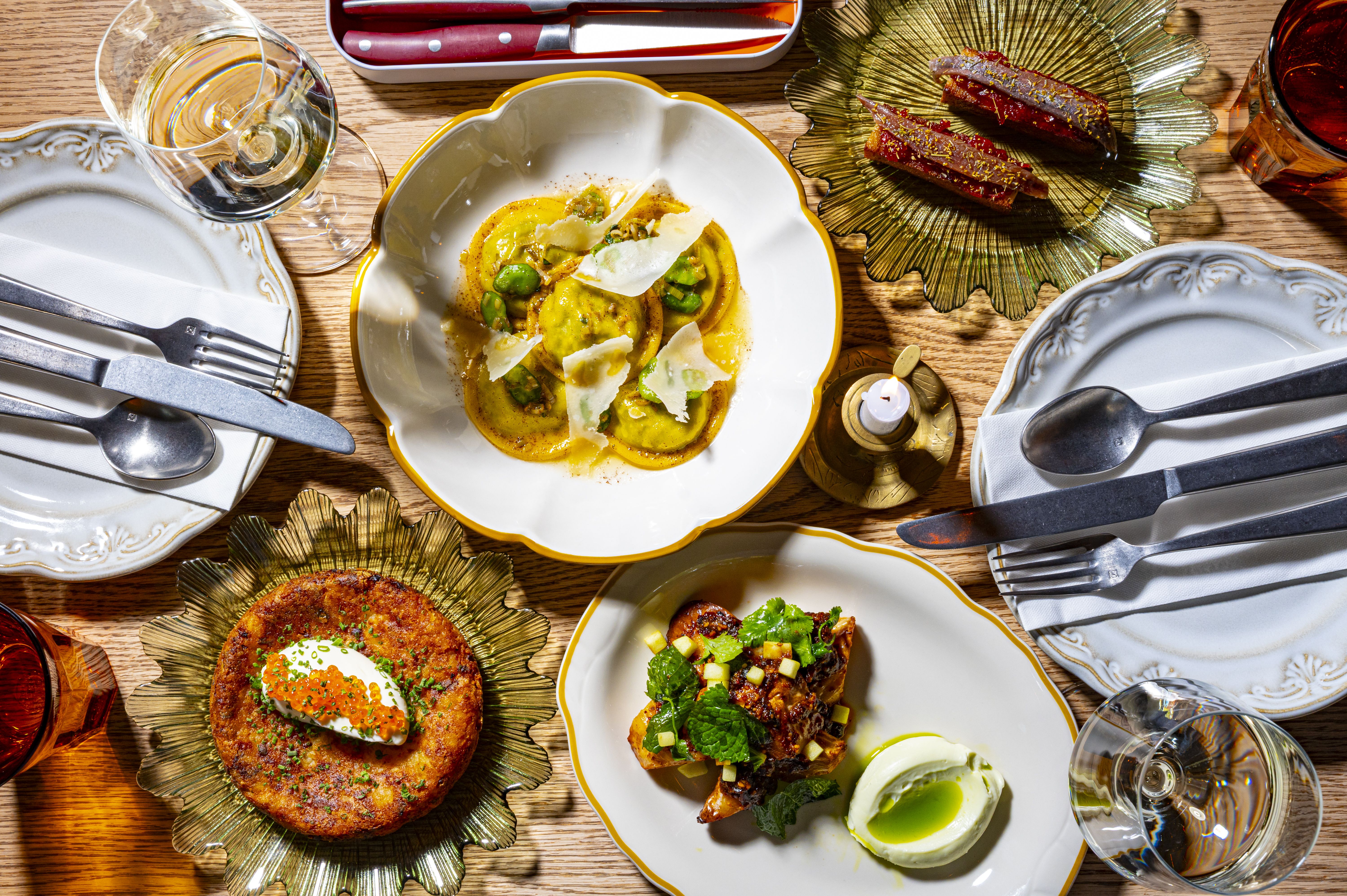Top-down view of a table with three dishes: center yellow-sauce ravioli with shaved cheese, bottom-left croquette on a gold leaf plate, and herb-topped meat on a white plate; glassware and cutlery.