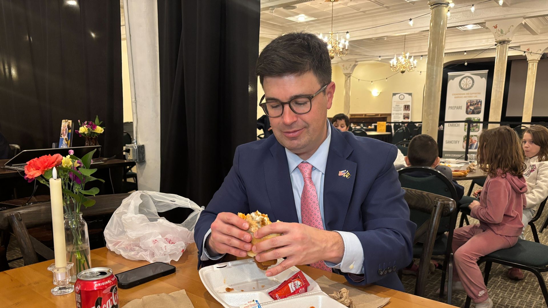 Man in blue suit and pink tie eating a sandwich at a wooden table with fries, coleslaw, ketchup, and a can of Coca-Cola in a room with chandeliers and children seated in the background.