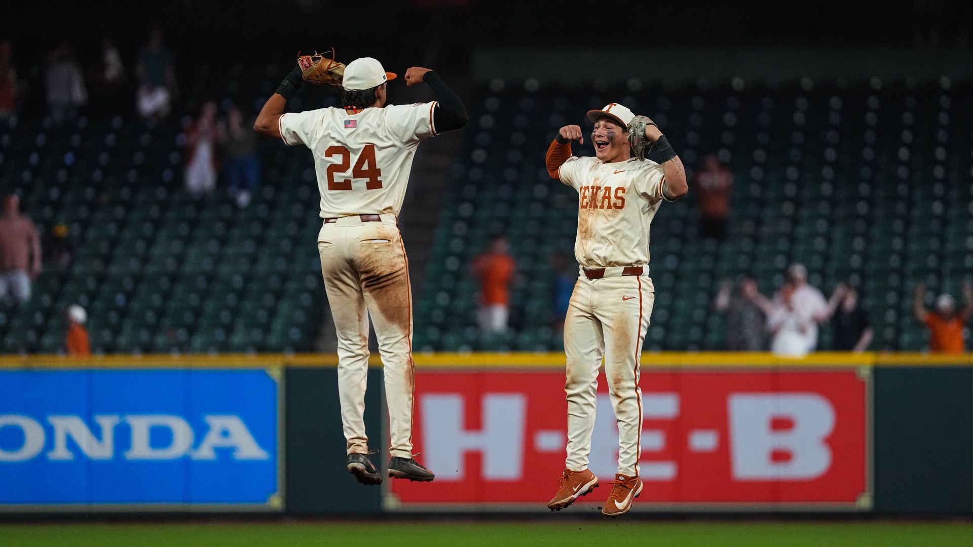 Two Texas baseball players in white and orange celebrate mid-air on the field; left player #24 with glove raised, right player with TEXAS on his chest smiles as empty stands loom behind.
