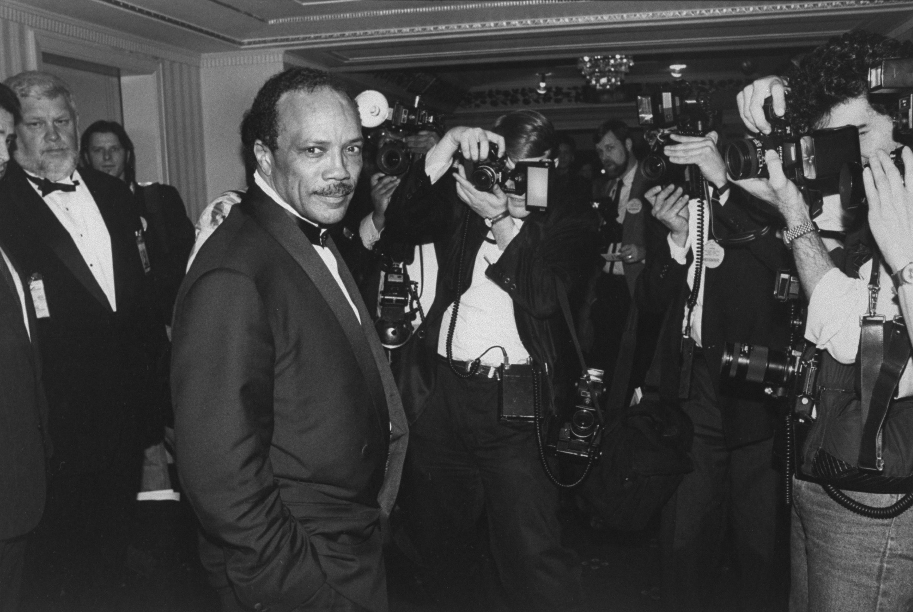 A black and white photo of musician Quincy Jones in a tux surrounded by photographers.