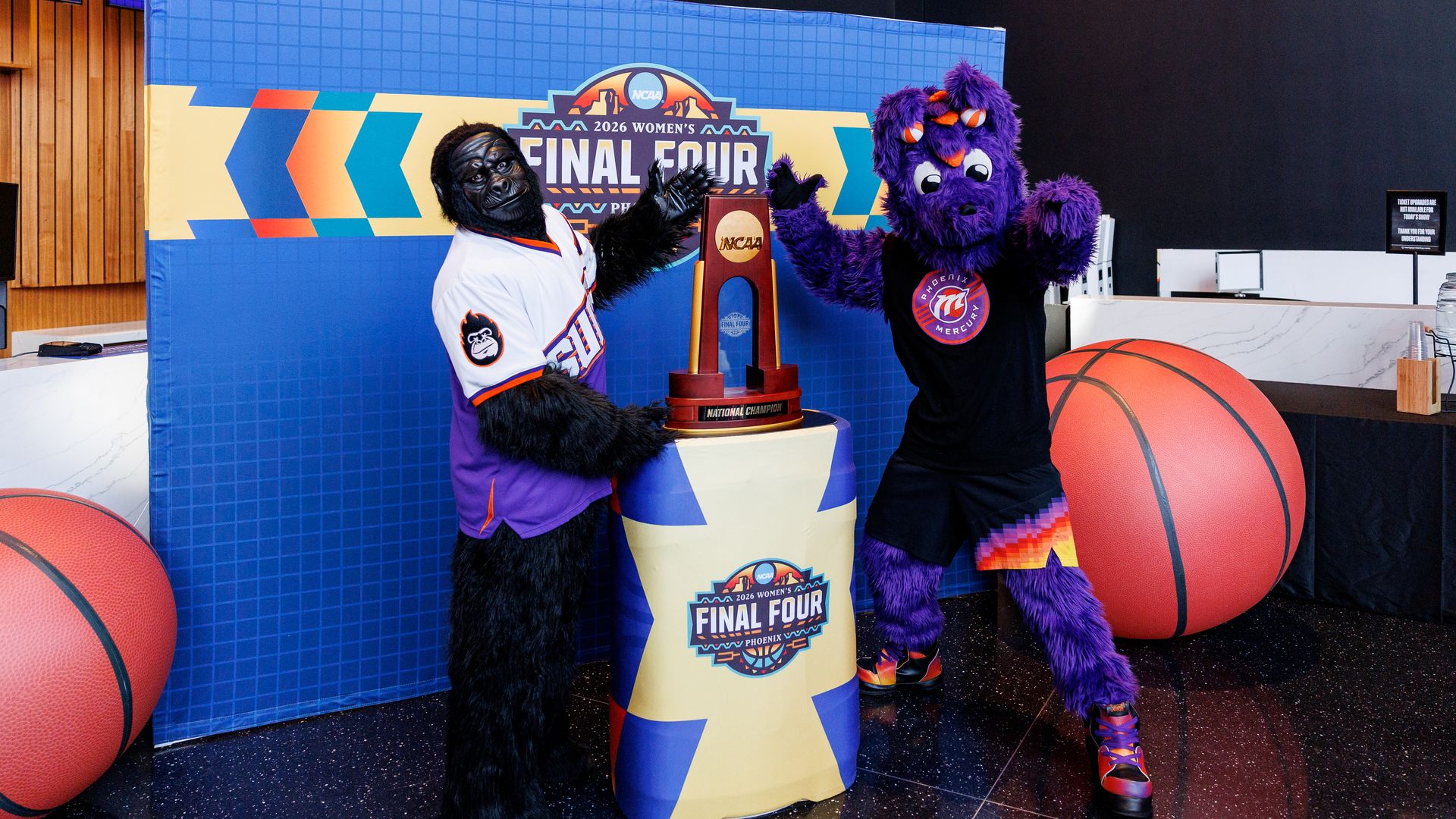Two colorful mascots, one gorilla and one purple creature, pose with the NCAA 2026 Women’s Final Four national champion trophy against a blue backdrop. Large basketball props flank them.