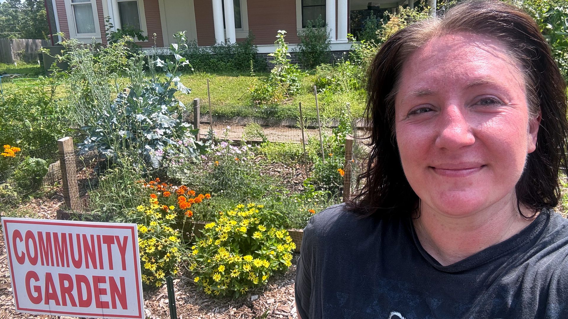 Smiling woman in a dark shirt takes a selfie in front of a pink house with white columns, beside a community garden with yellow and orange flowers and a sign.