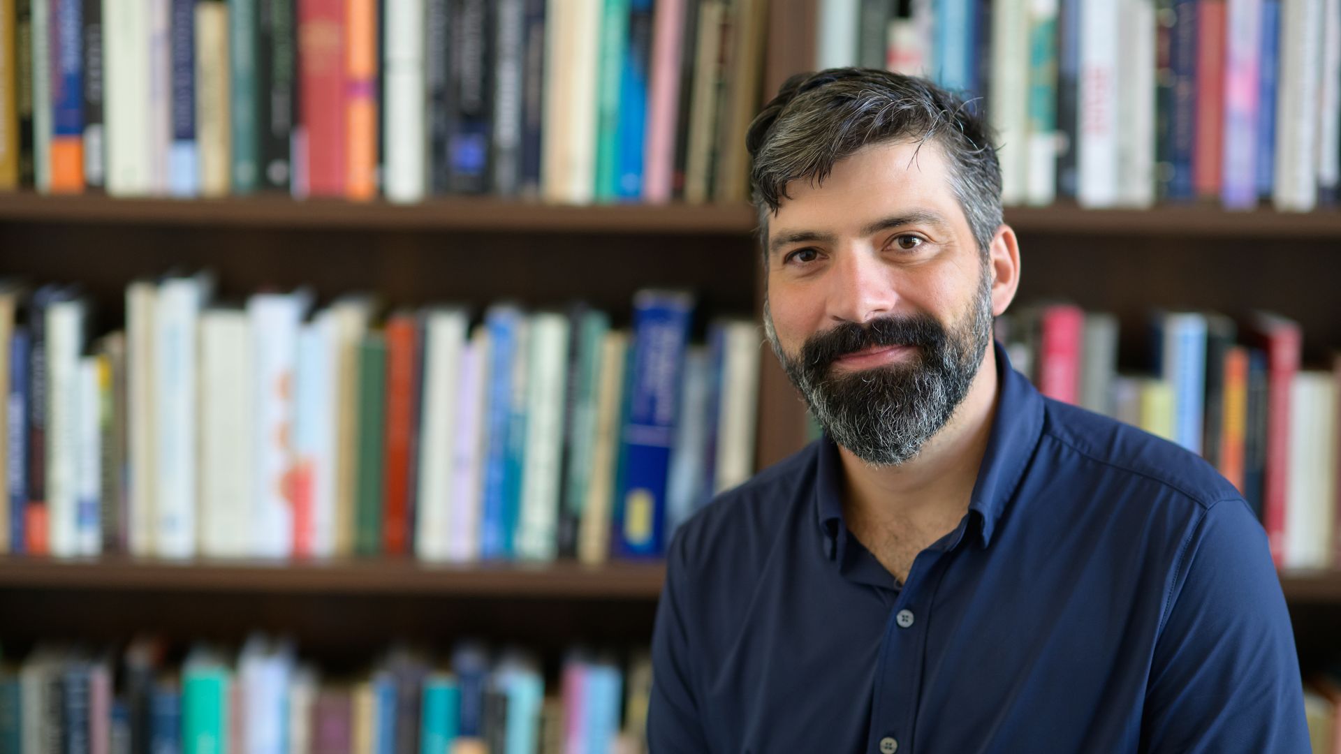 Portrait of a man with gray-streaked hair and a beard, wearing a dark blue shirt, smiling slightly, with a blurred bookshelf of colorful books in the background.