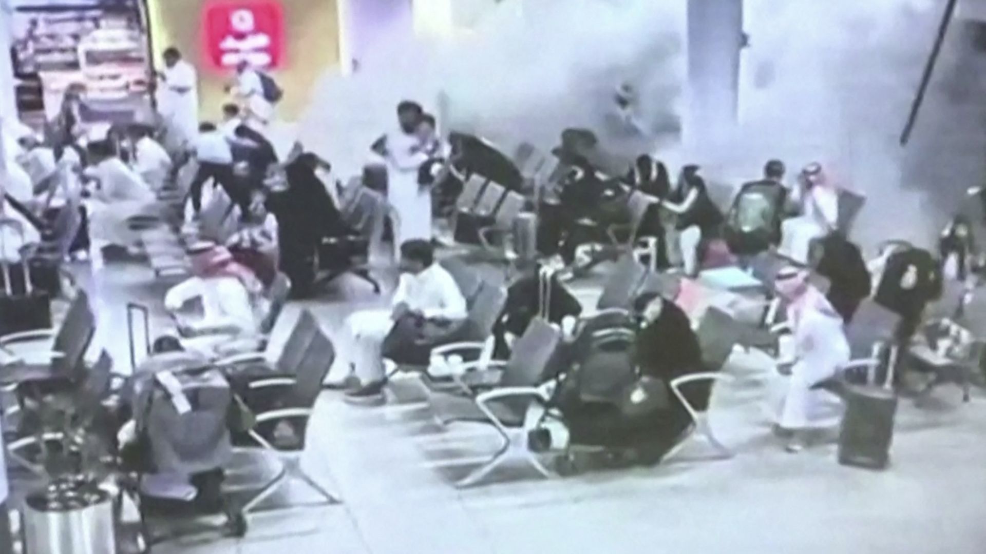 Crowded indoor dining area with dozens of people seated at scattered tables and chairs; a red sign on a yellow wall in the background, and a hazy, busy scene with a tall pillar on the right.