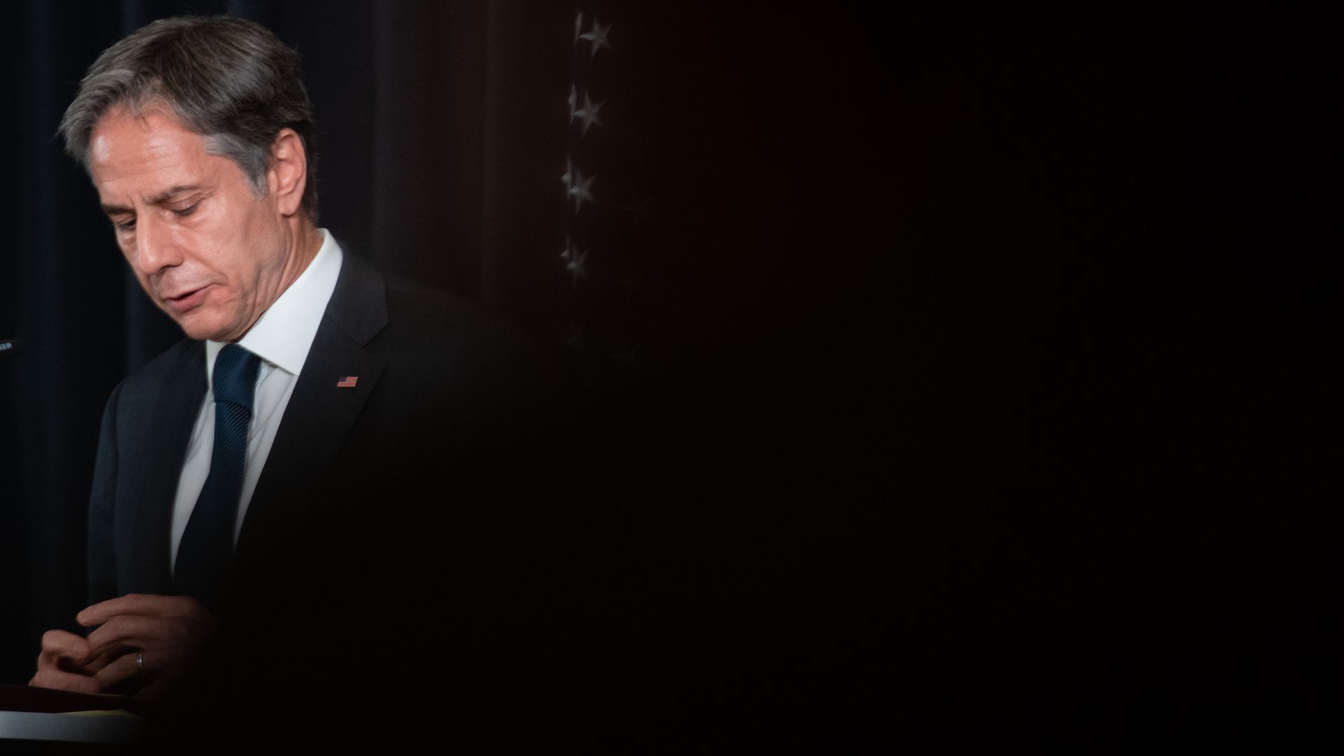 Secretary of State Tony Blinken looks down before speaking agains a dark curtained backdrop