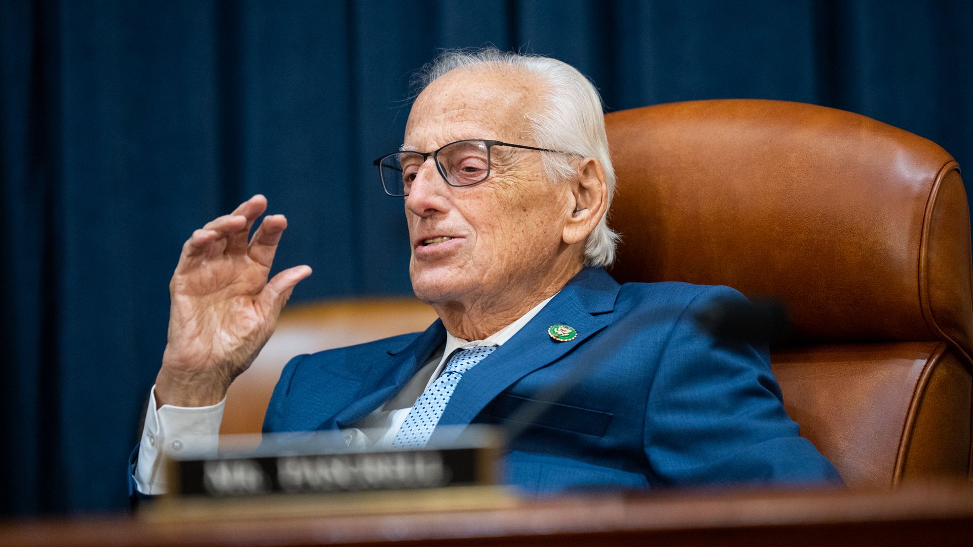 Rep. Bill Pascrell, wearing a blue suit, white shirt and blue tie, sitting in a brown leather committee room chair in front of a blurred plaque.