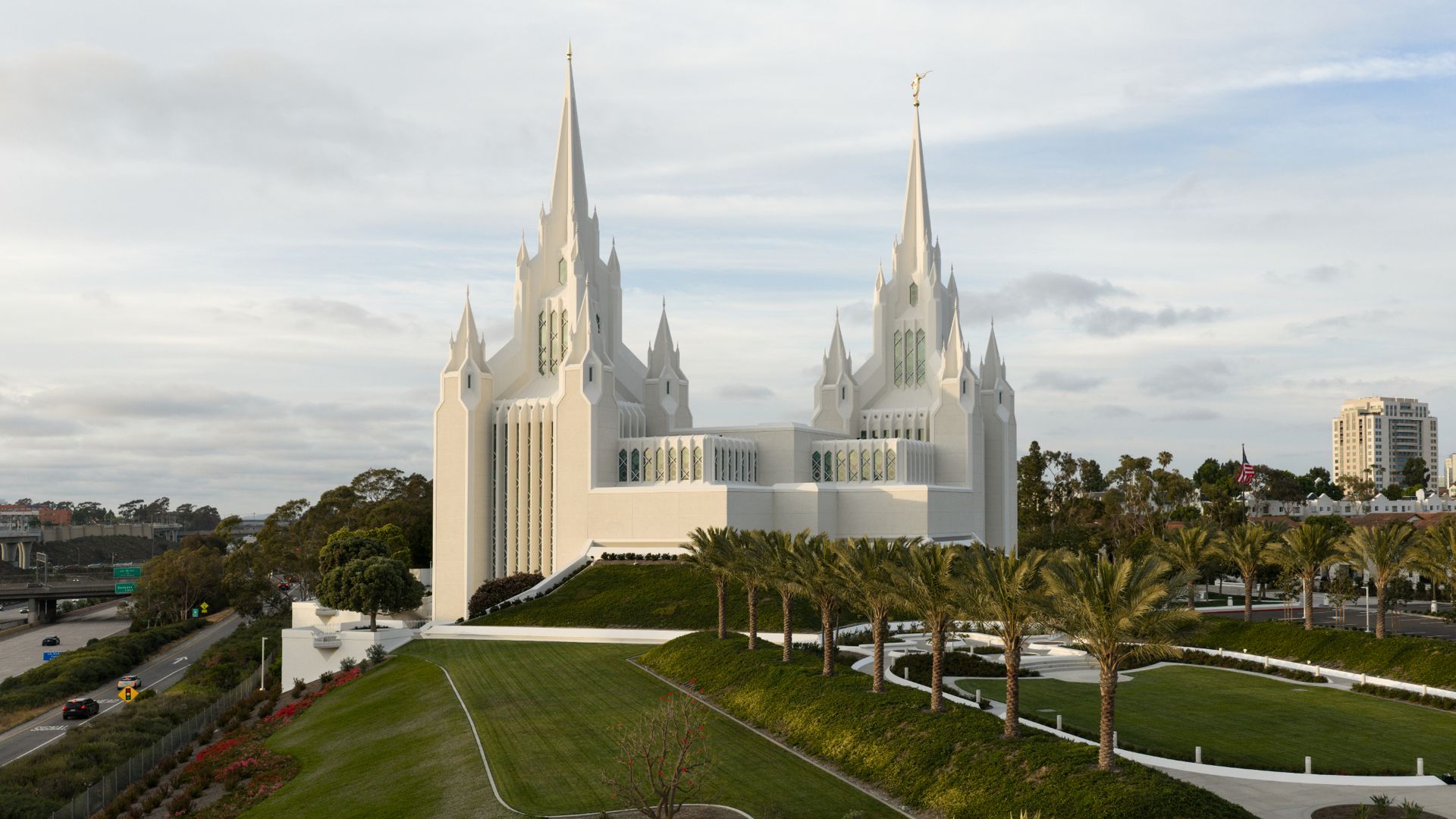 White twin-spired Mormon temple with tall pointed spires topped by statues, sits on a manicured lawn with palm trees; blue sky, distant city skyline, and a freeway nearby.