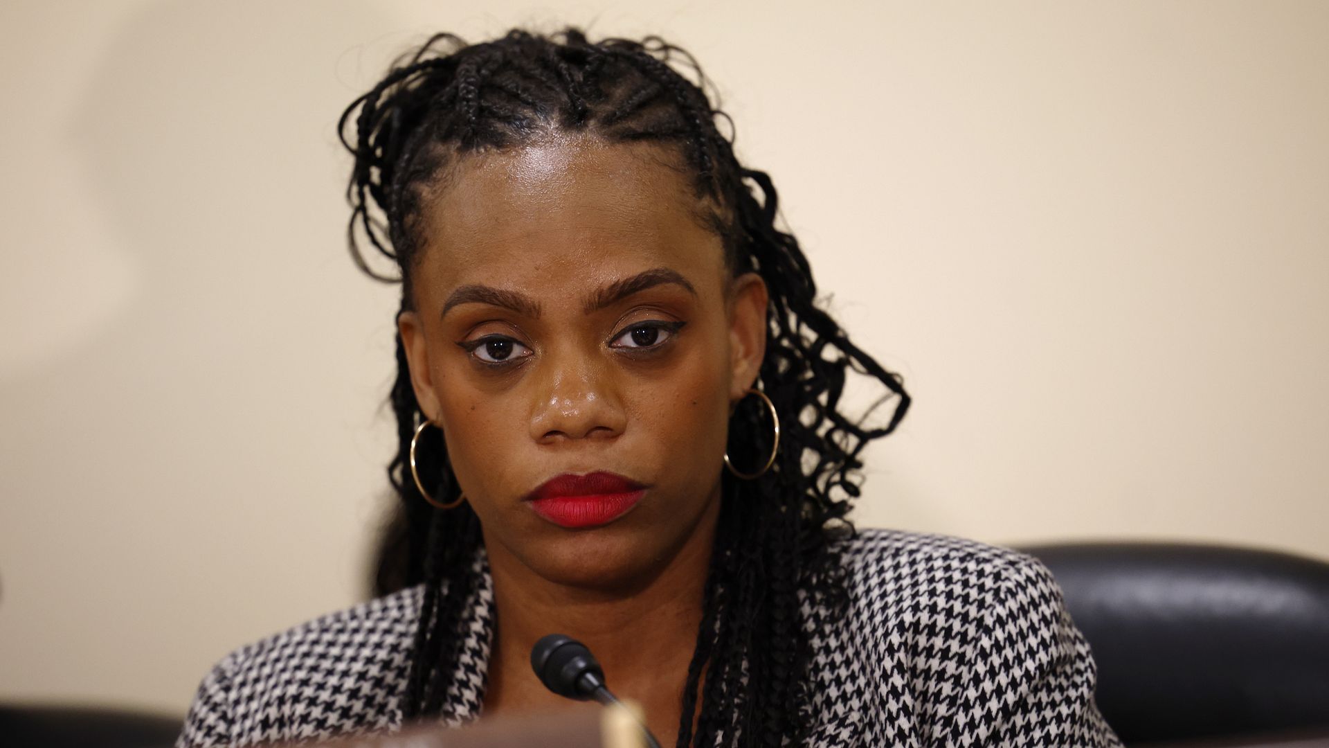 Close-up of a U.S. Rep. Summer Lee, wearing hoop earrings, red lipstick, and a black-and-white houndstooth blazer, seated in front of a microphone and nameplate.