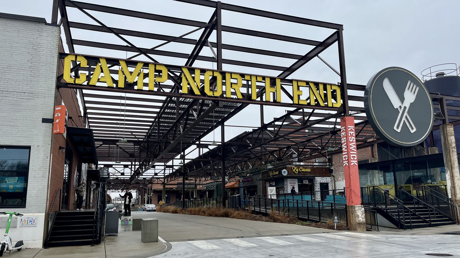 Entrance to Camp North End with large yellow sign on black steel frame, gray sky, pedestrian crossing, and nearby buildings including a restaurant with a knife and fork logo.