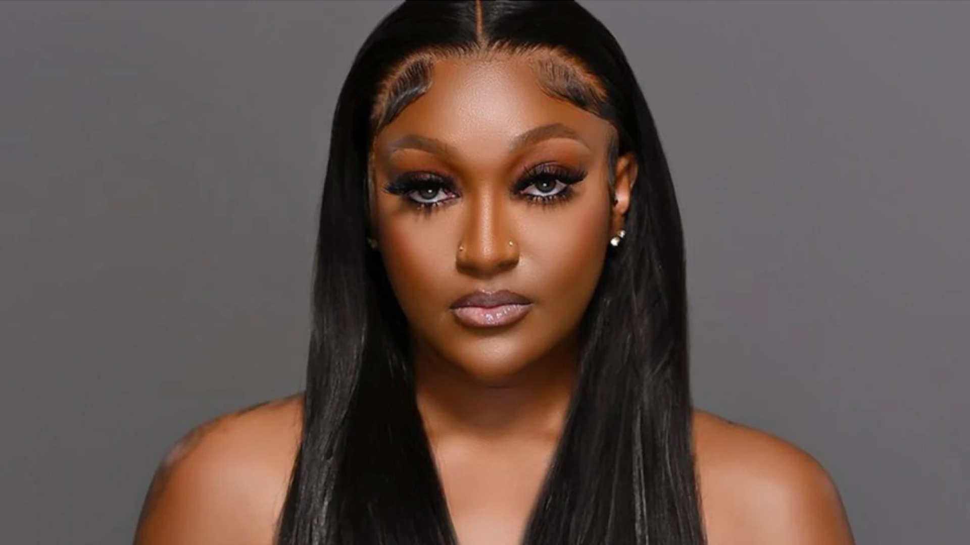 Portrait of a woman with long straight black hair, brown skin, dramatic eyelashes, subtle makeup, and nose piercings, against a plain gray background.