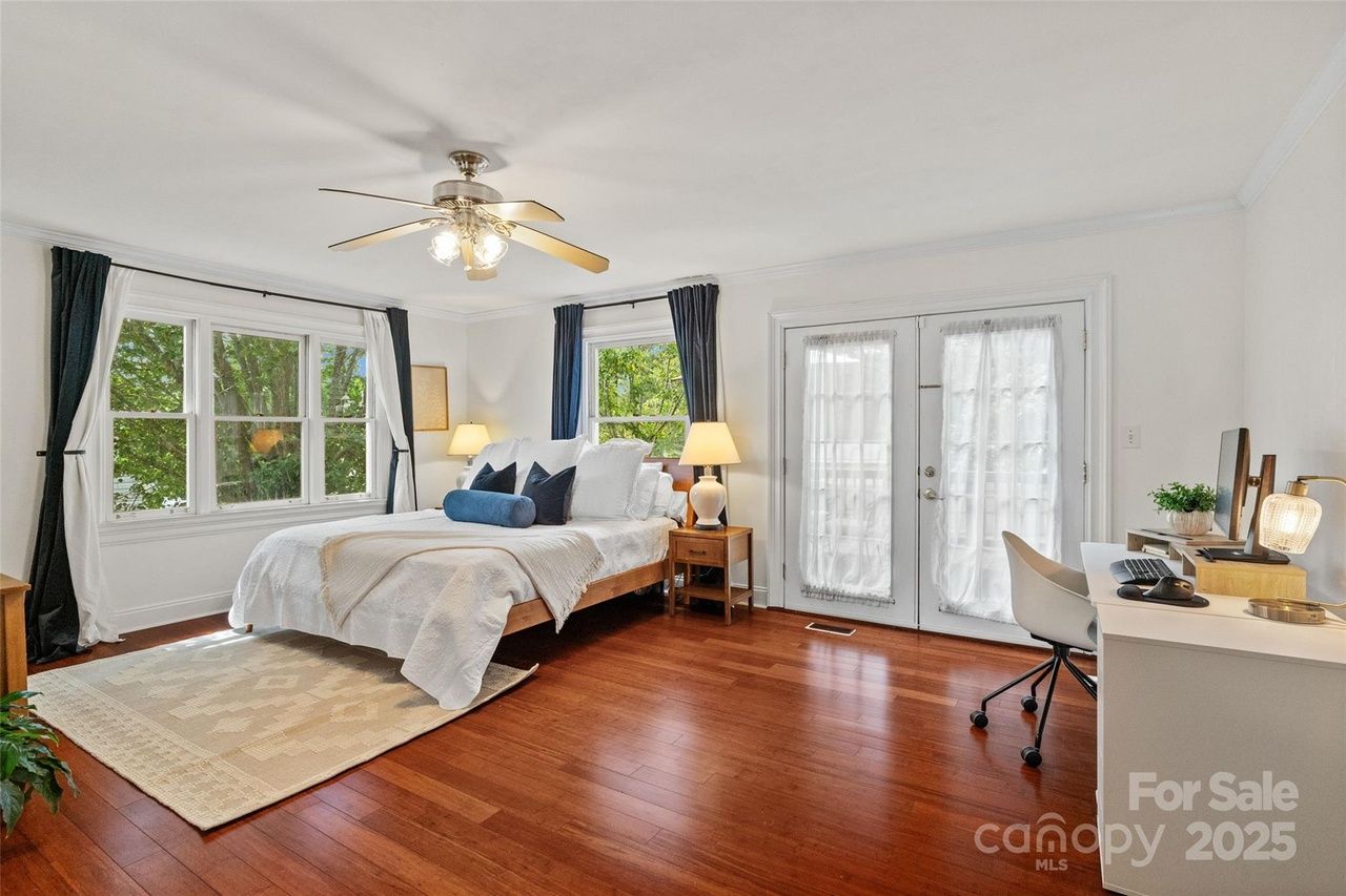Bright bedroom with white walls, wooden floor, queen bed with white and blue bedding, two bedside lamps, ceiling fan, large windows with dark curtains, French doors, and a desk with chair and computer.