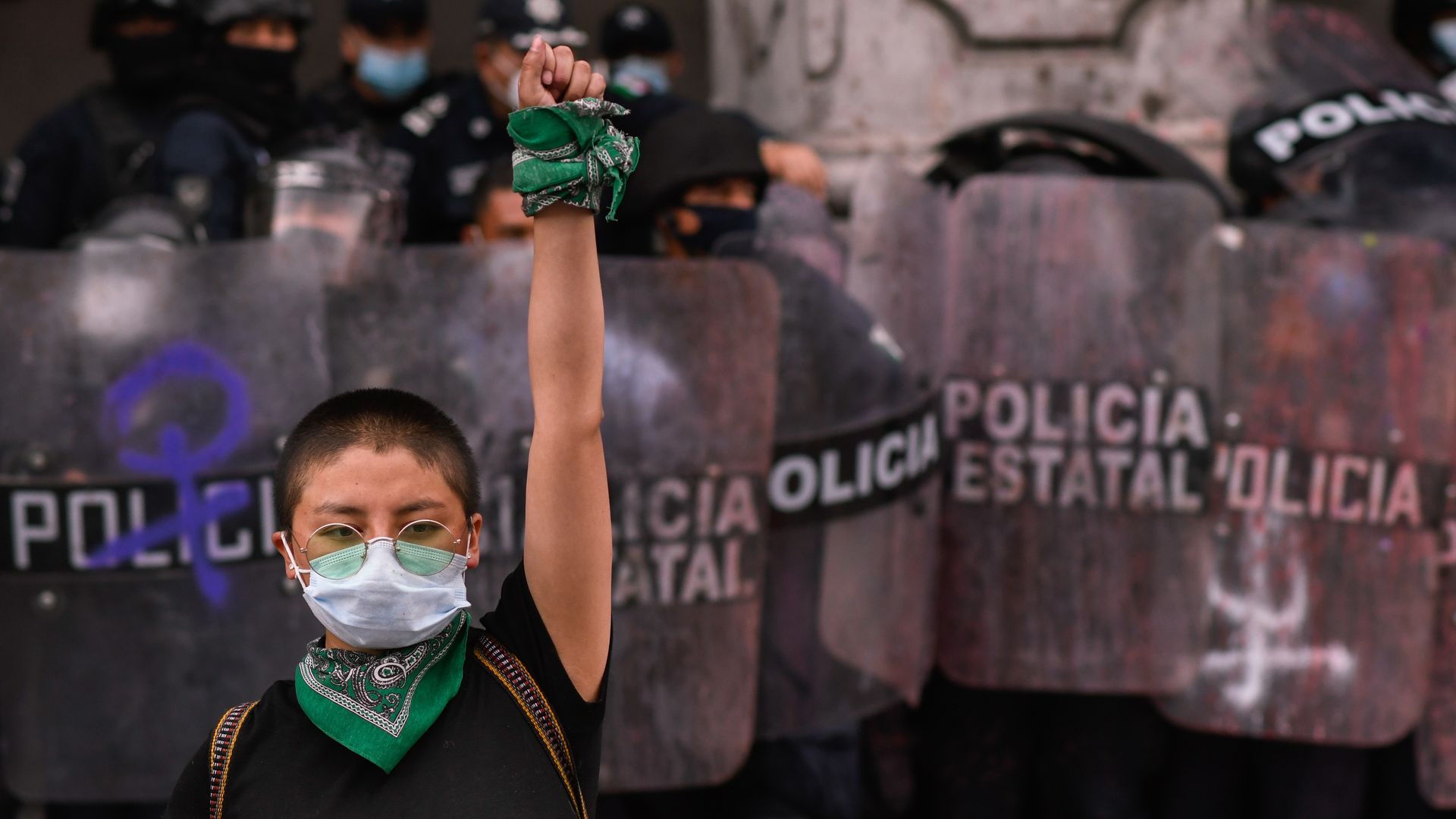 Photo of a masked protester raising their arm with police in the backdrop