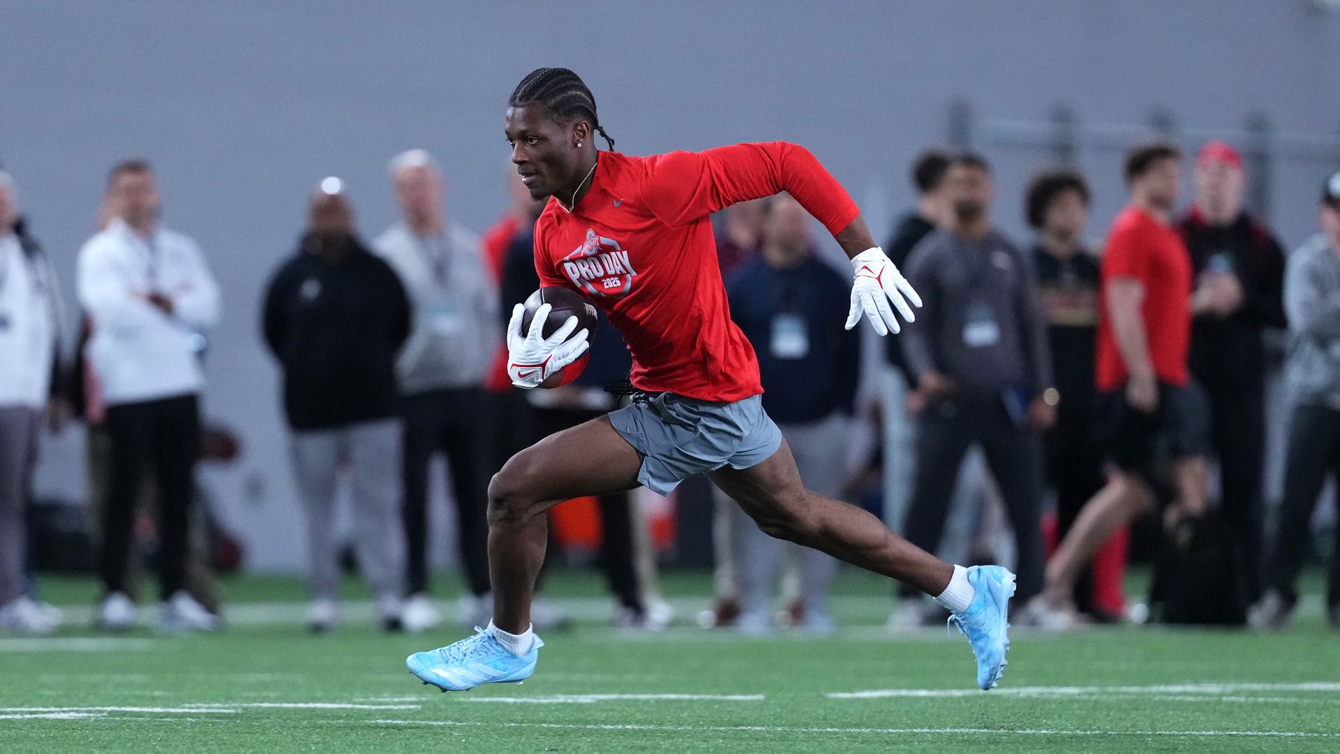 Carnell Tate, a young man wearing a long-sleeved red shirt and gray shorts, runs across a football field with a ball tucked in his right arm as dozens of scouts watch in the background.