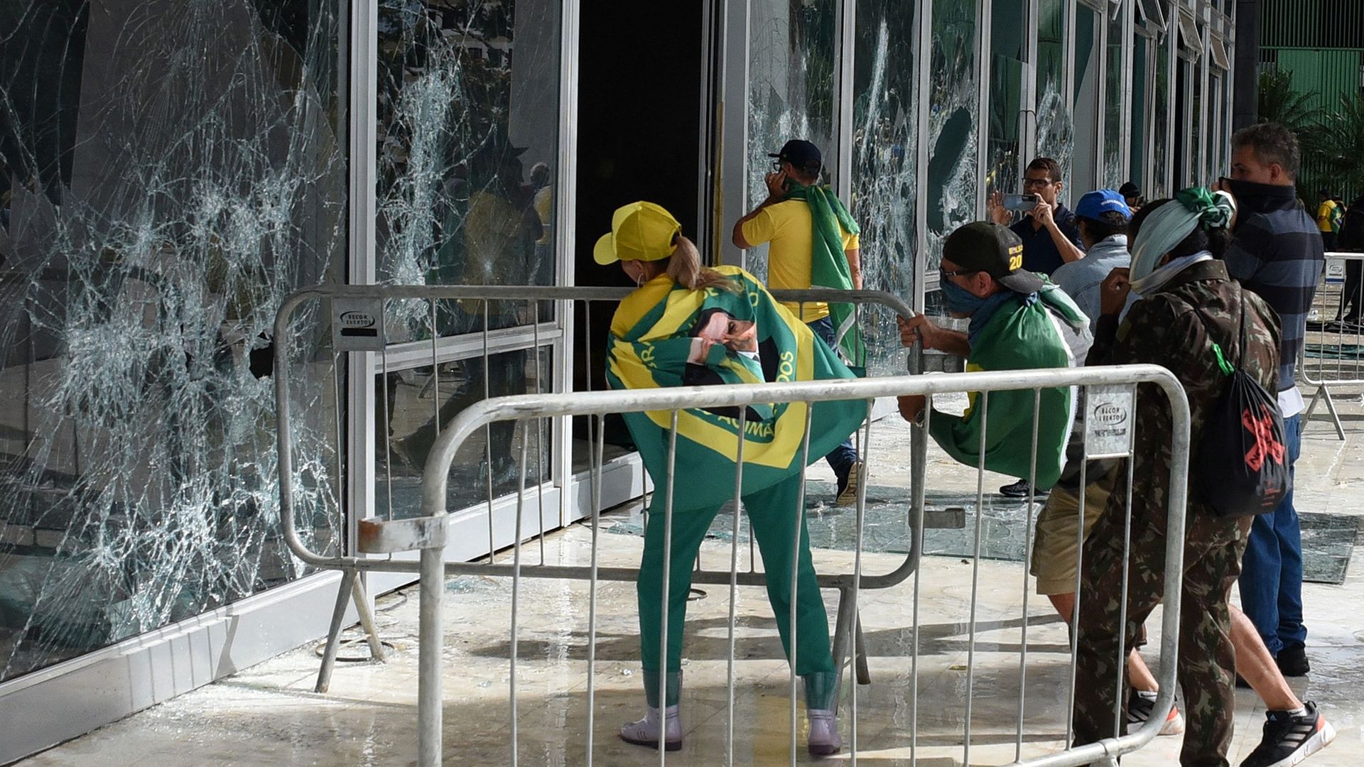 rioters wearing brazilian flags on their backs break large glass windows at government buildings