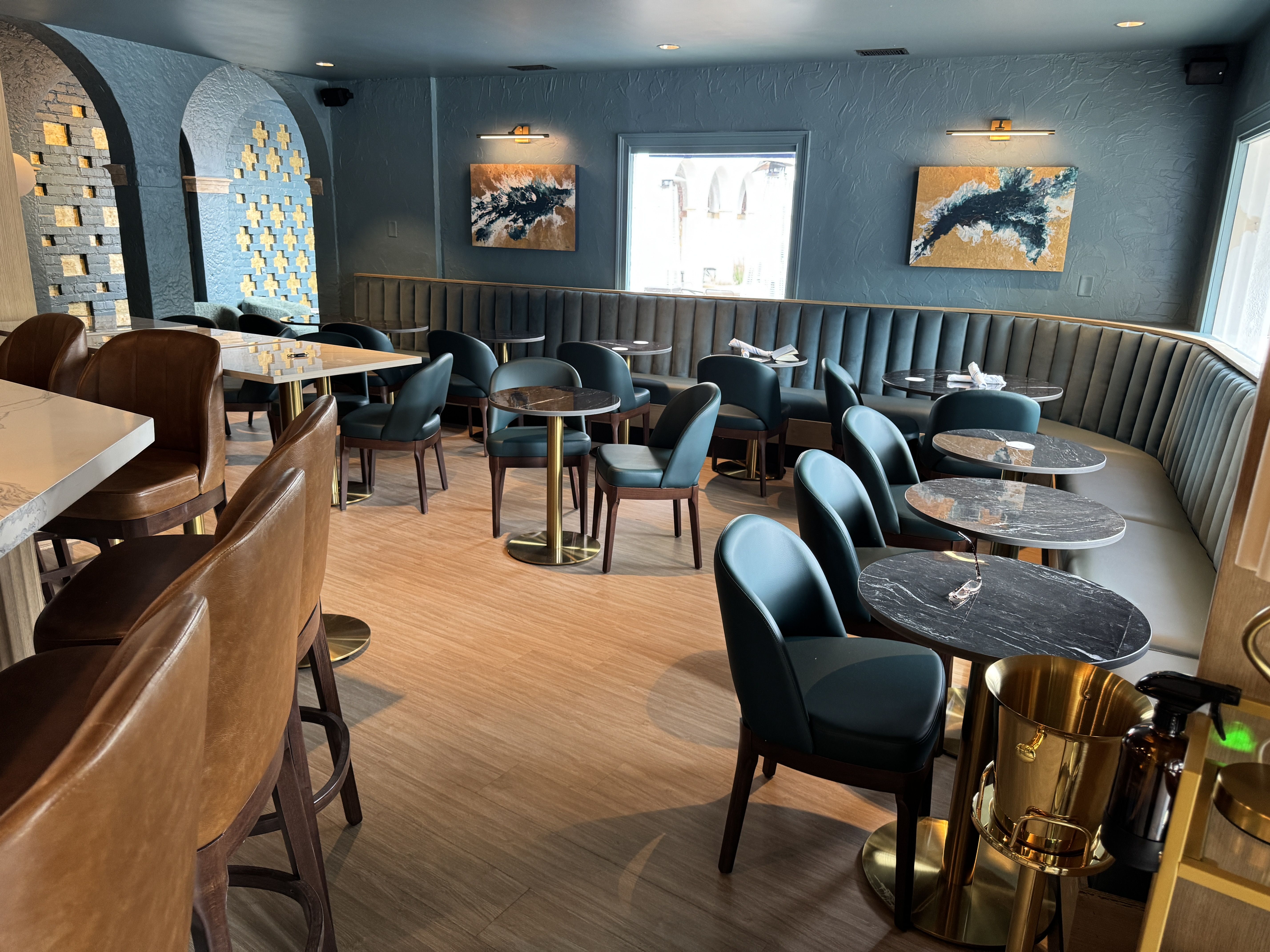 Modern café interior with teal walls, brown leather bar stools, teal chairs, black marble tables with gold bases, arched wall accents, abstract paintings, and wood flooring.