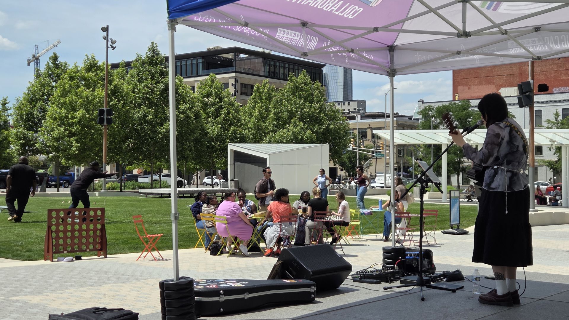Outdoor stage under a purple canopy; a female guitarist sings into a mic while a diverse audience sits at colorful chairs around tables, with trees and city buildings in the background.