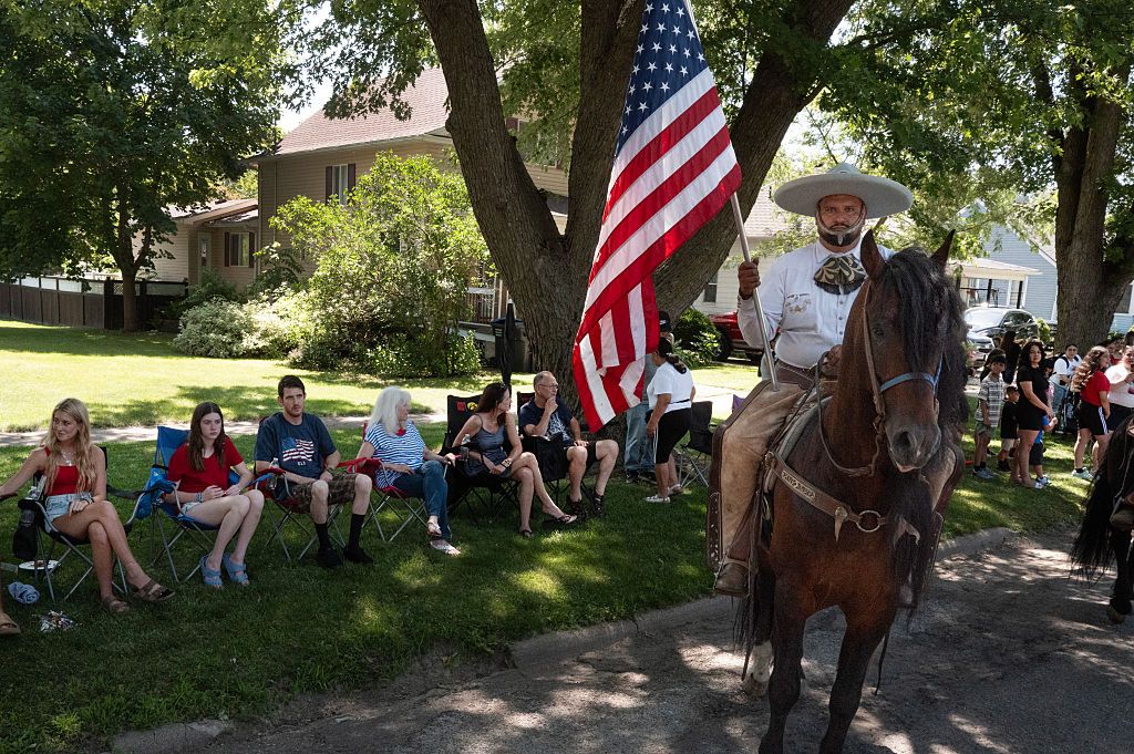 A photo showing a man wearing a wide brimmed, sombrero-like hat, holding a large flag and riding a horse. Onlookers sit in lawn chairs in grass.