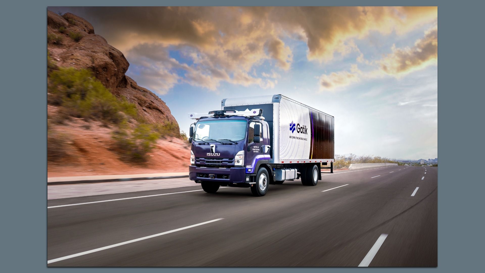 A purple and white Isuzu truck equipped with Gatik's driverless technology drives on a highway beside desert terrain under a partly cloudy sky at sunset.
