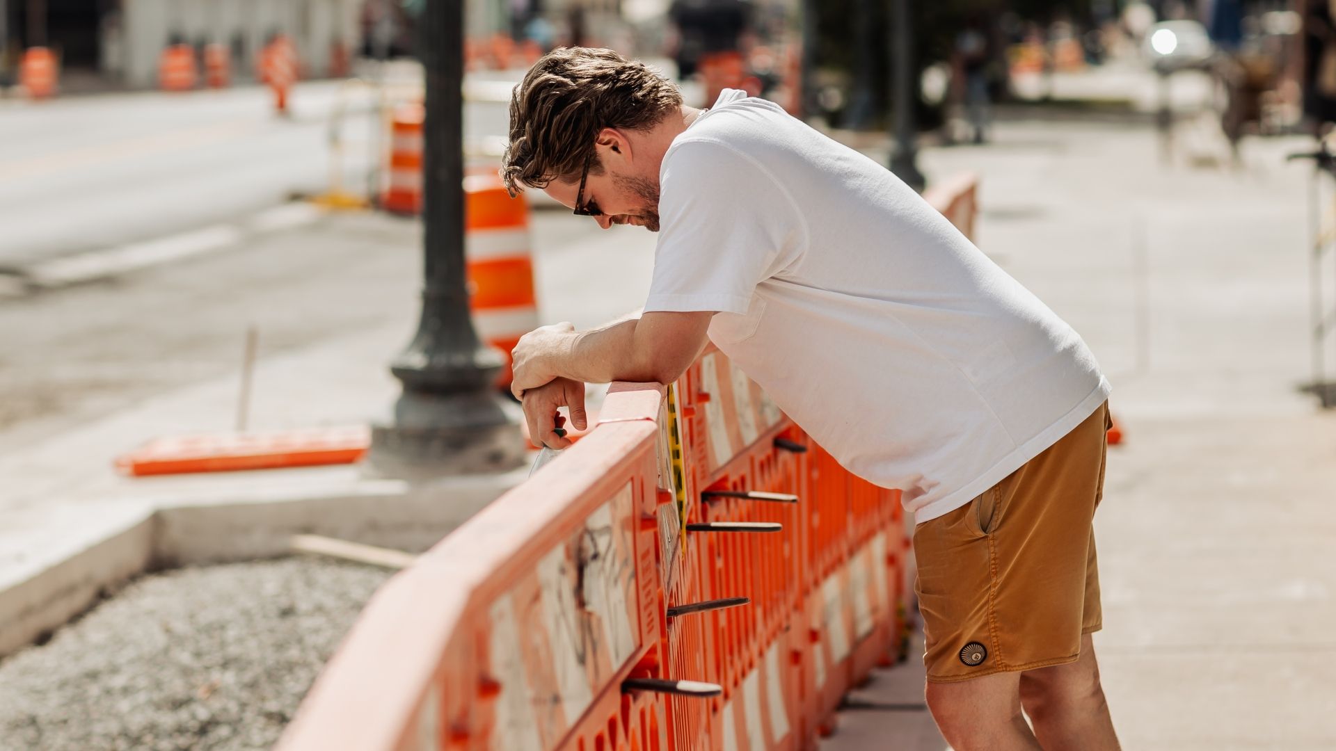 A man in a white T-shirt and tan shorts leans on an orange construction barrier on a sunny city street, looking down with sunglasses; blurred urban background.