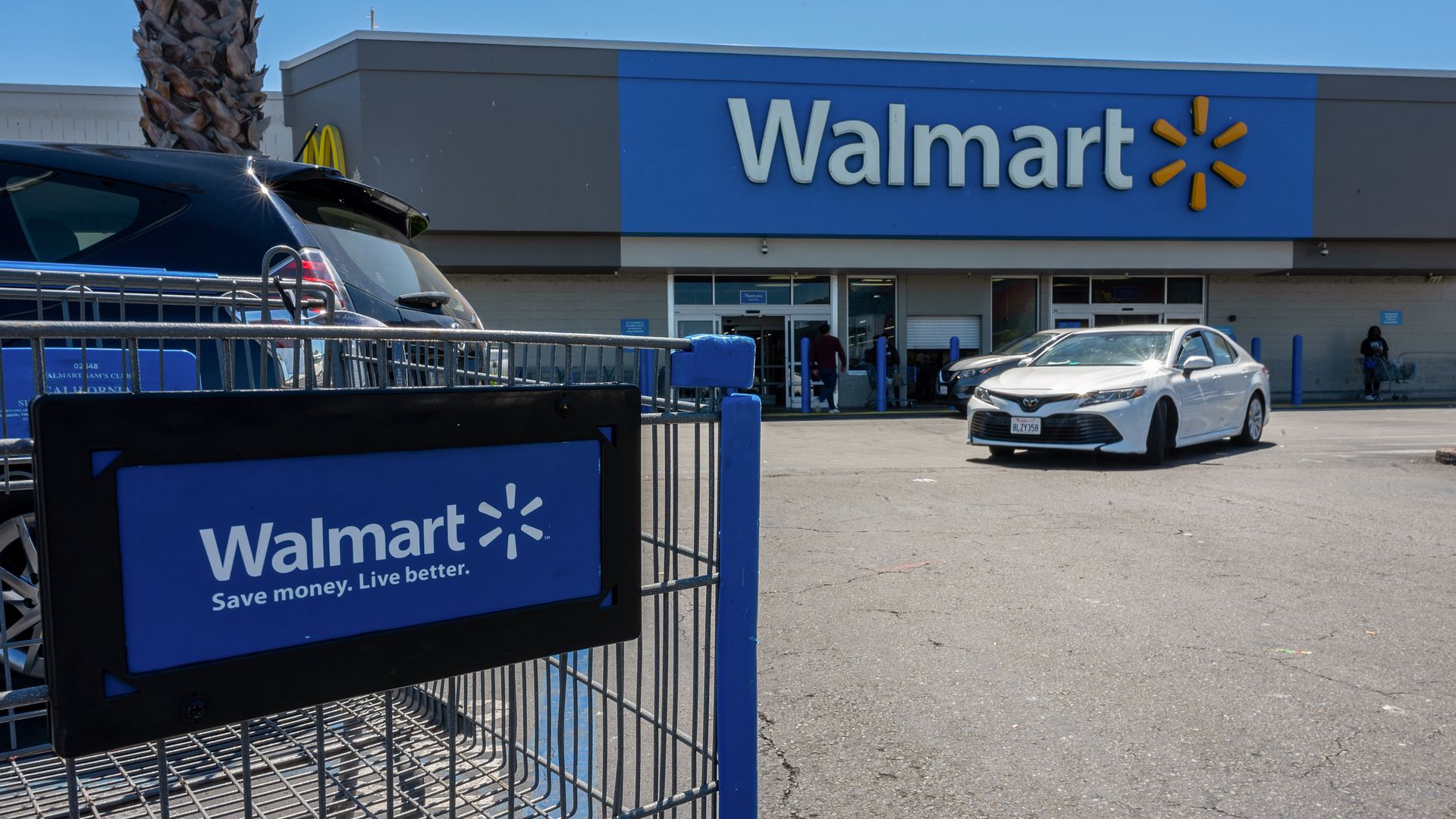 Shopping cart with blue Walmart sign and slogan "Save money. Live better." in front of a Walmart store.