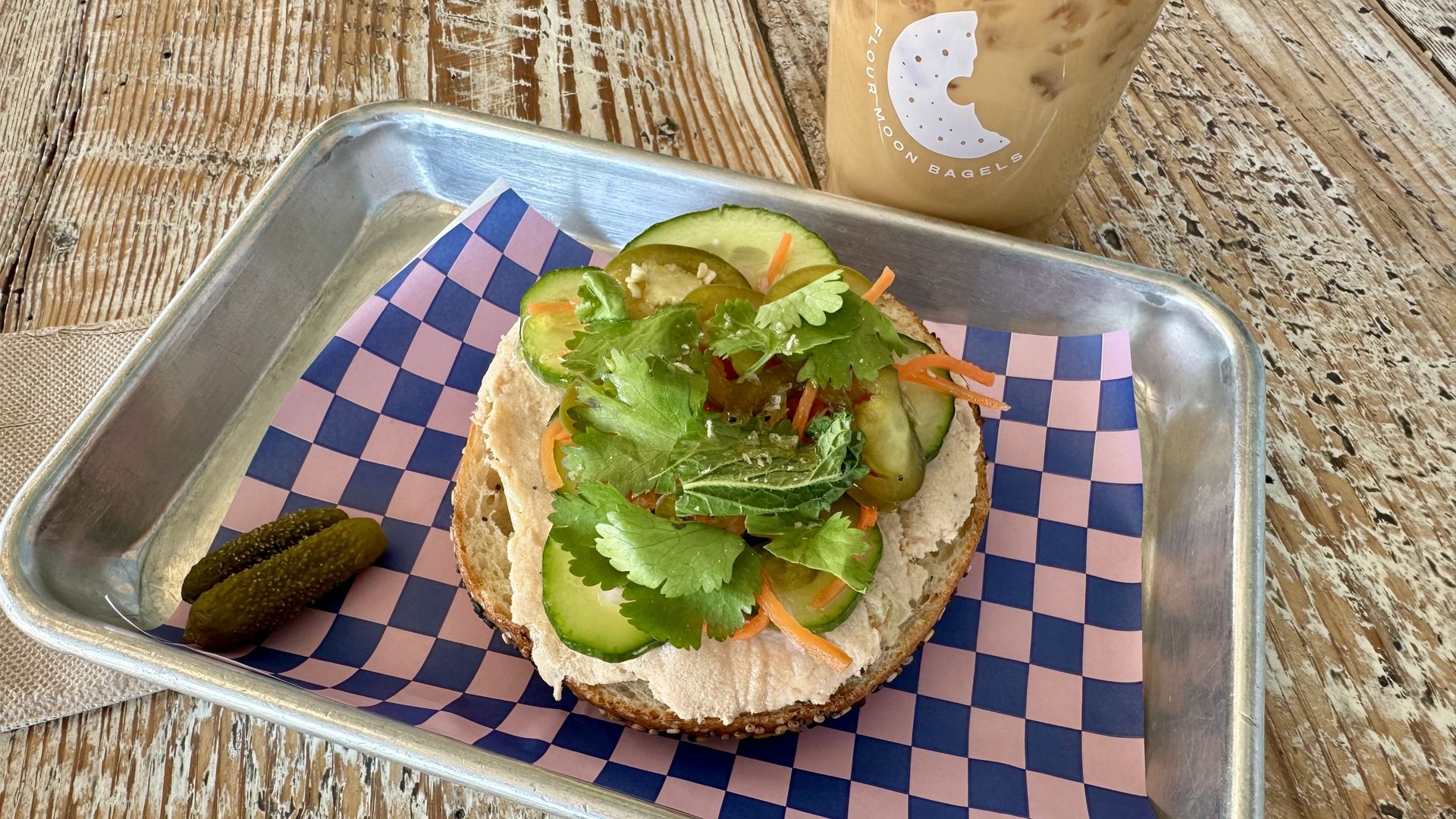 Open bagel sandwich with salmon pate, cucumber slices, pickles, shredded carrots, and cilantro on blue and pink checkered paper in a metal tray, with iced coffee in the background.
