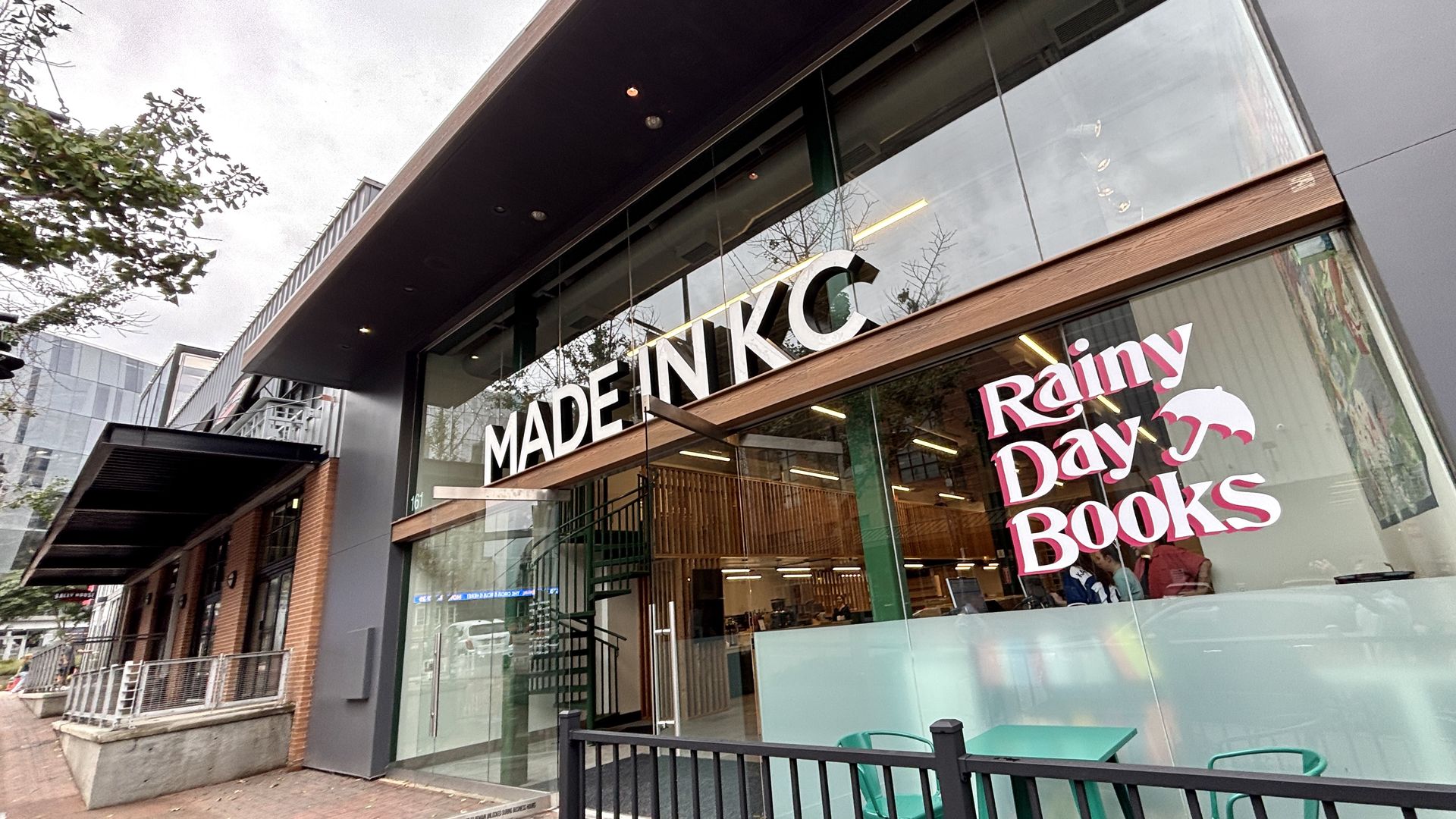 Angled view of a bookstore storefront with glass windows and wooden trim. Large white letters spell "MADE IN KC" across the window; a pink "Rainy Day Books" sign with an umbrella is visible inside.