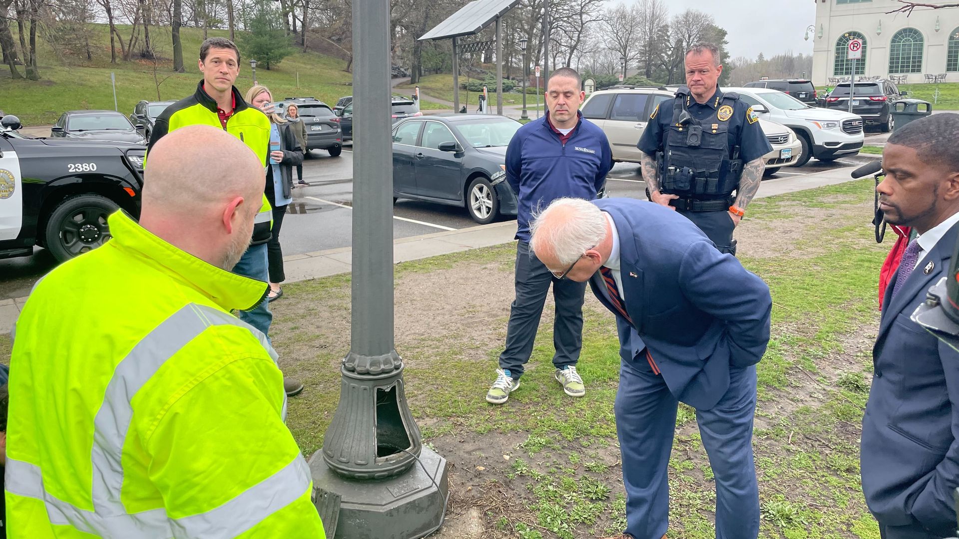 TIm Walz looks at the bottom of a light post surrounded by Mayor Melvin Carter, Police Chief Todd Axtell and city crews