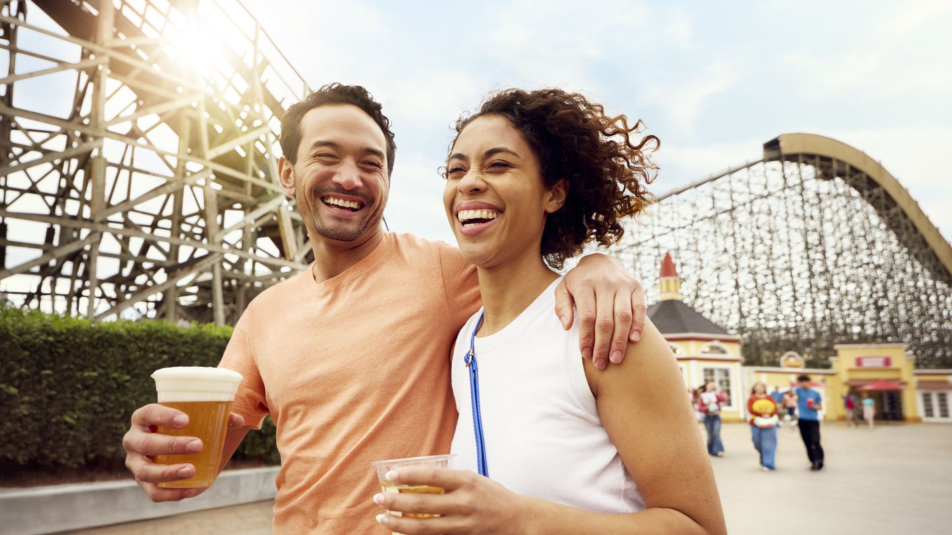 Smiling couple holding drinks with wooden roller coaster and amusement park buildings in the background on a sunny day.
