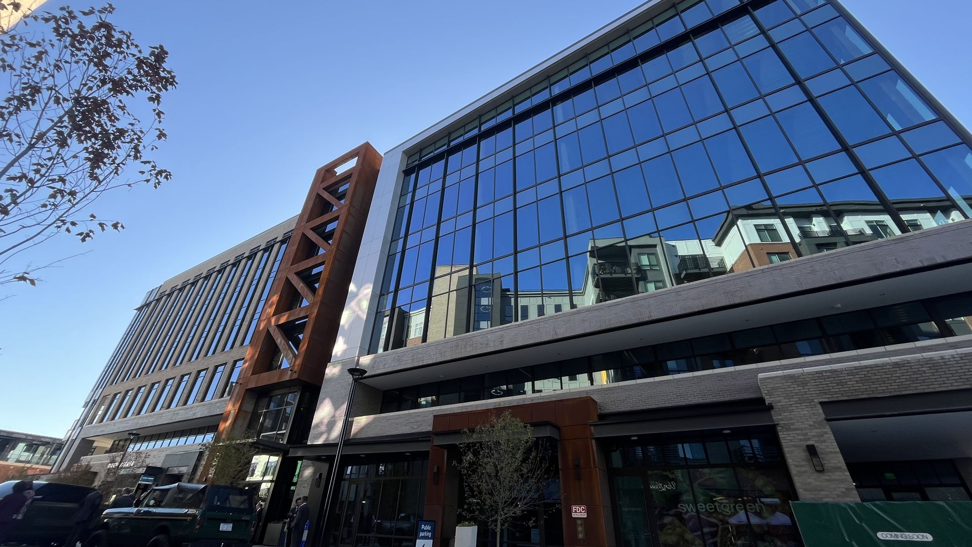 Modern multi-story building with large reflective glass windows, rust-colored metal accents, a few parked vehicles, young trees, and a clear blue sky.