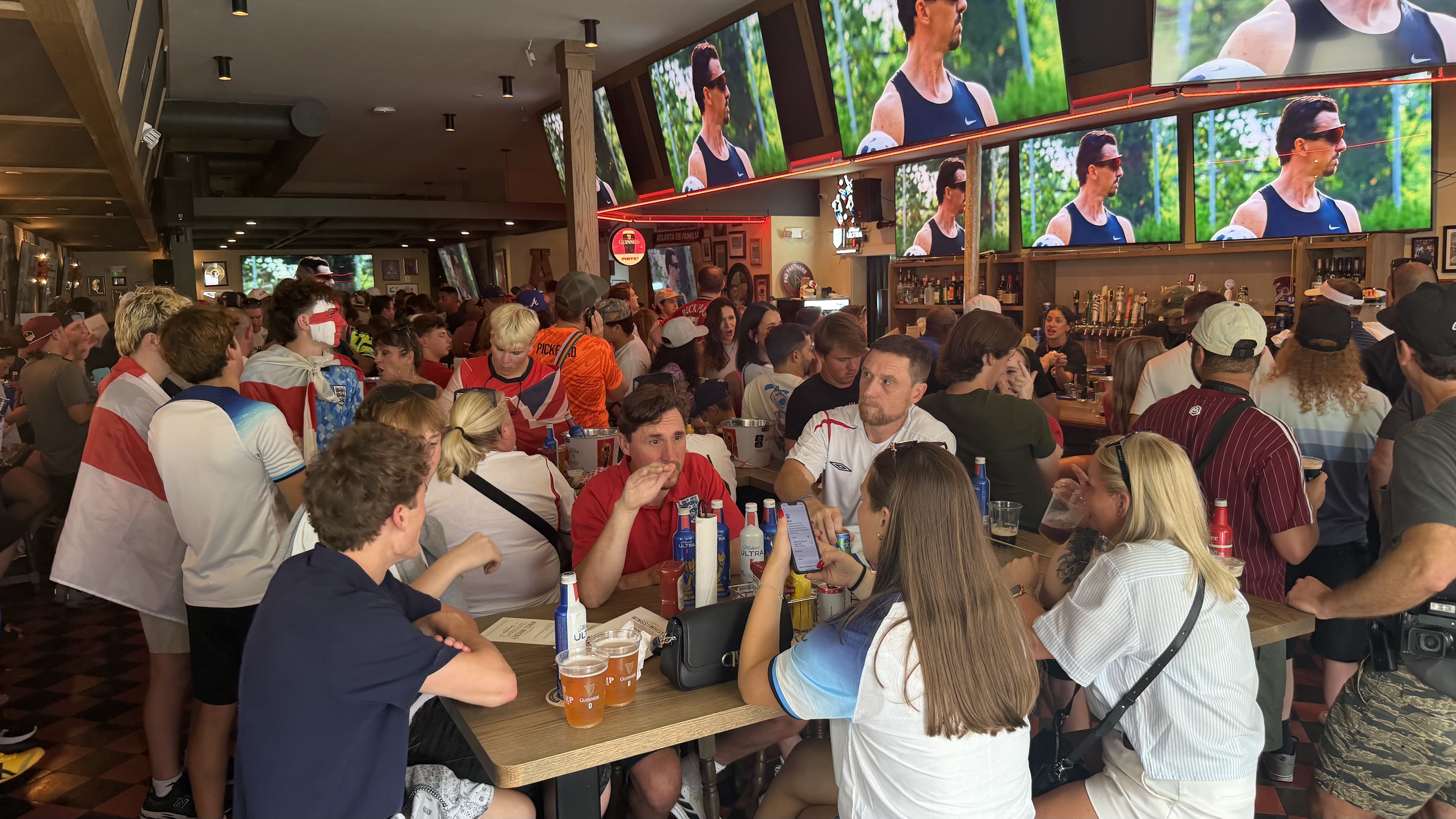 Busy sports bar full of fans in jerseys, crowded tables with drinks, and a back bar. Large screens show a man in a black tank top running outdoors.