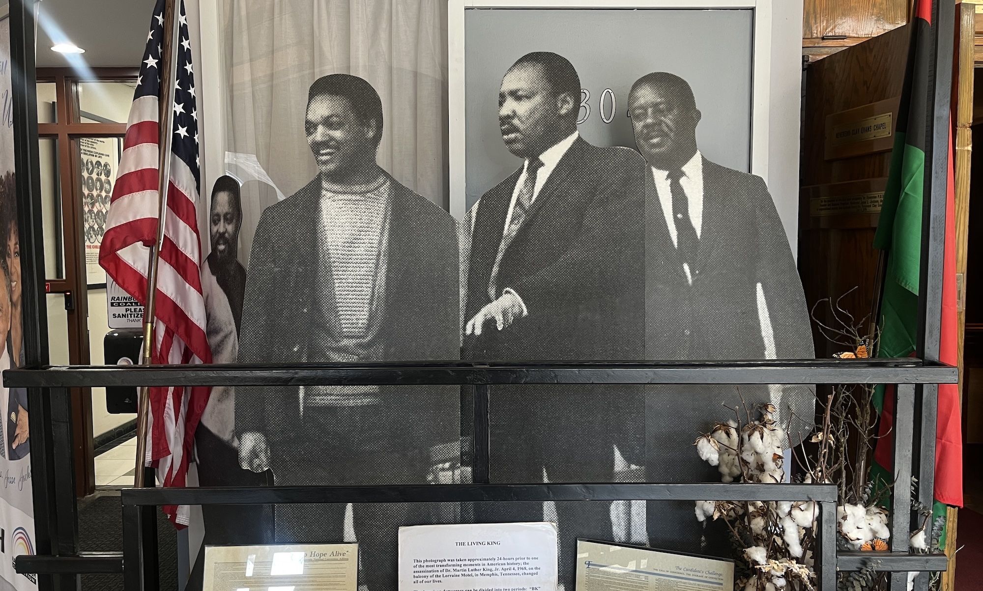 Black and white cutouts of Jesse Jackson, Jr, Martin Luther King and Ralph Abernathy in suits and sweaters displayed indoors between American and Pan-African flags, with cotton stalks and informational plaques below.