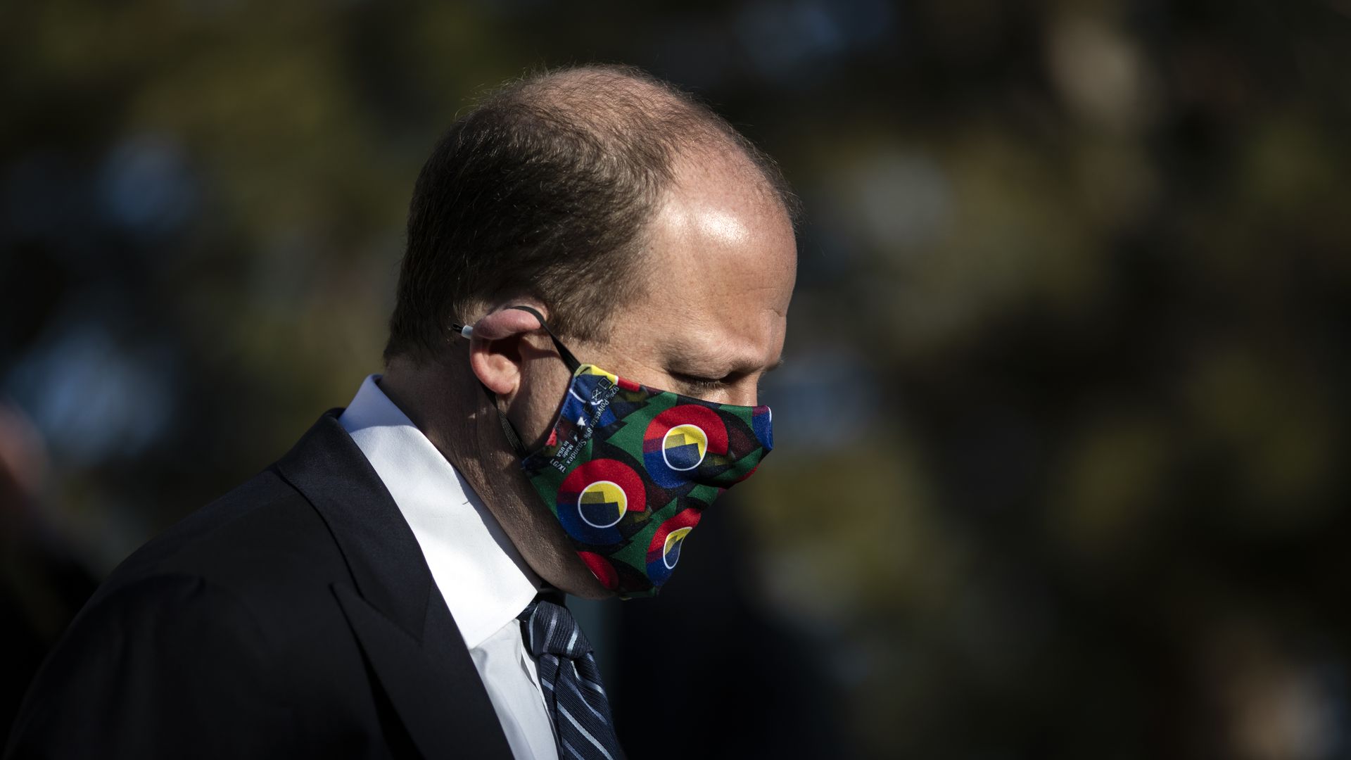 Photo of Jared Polis in a black suit and a face mask that displays the Colorado seal