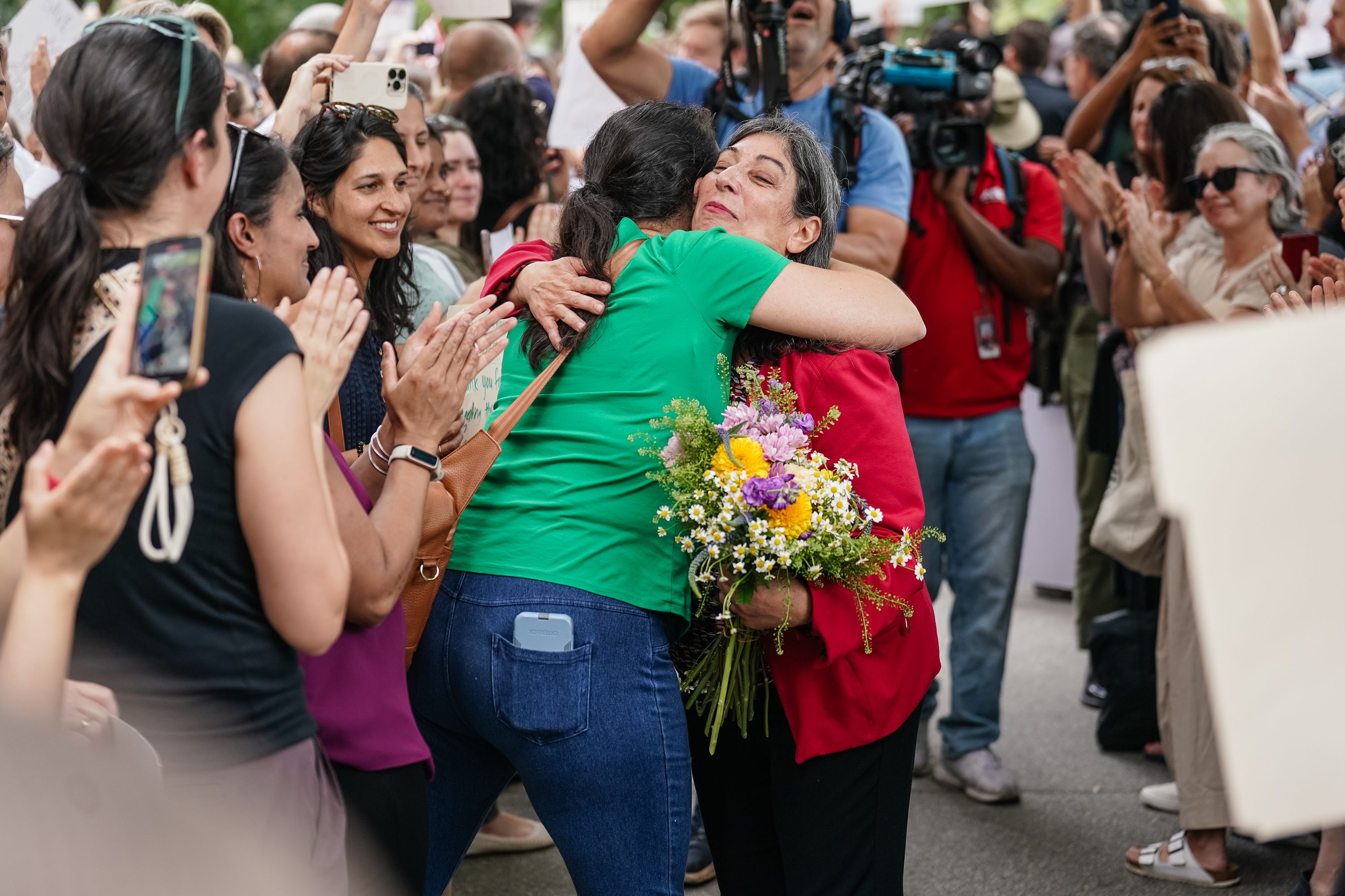 Two women hugging, one in a green shirt and jeans, the other in a red jacket holding a colorful bouquet, surrounded by applauding people and photographers at an outdoor event.