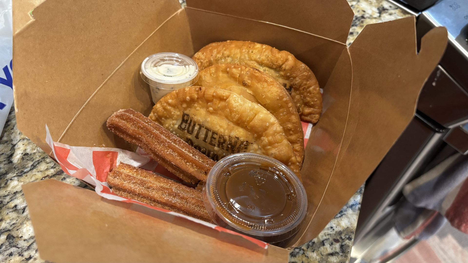 Open brown takeout box containing golden-brown pastries (rounds and a churro-like stick) with small cups of dipping sauces on a speckled granite counter.