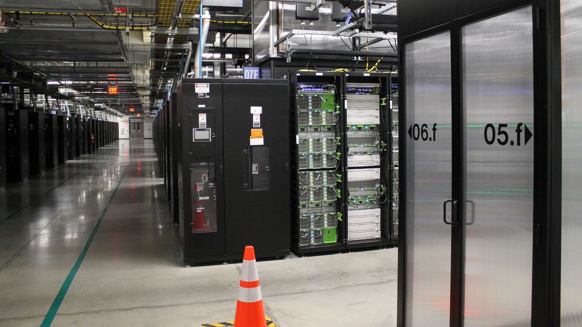 Industrial data center aisle with tall black server racks on both sides, yellow overhead cable trays, and a glass-front cabinet on the right; an orange traffic cone sits on the polished floor.
