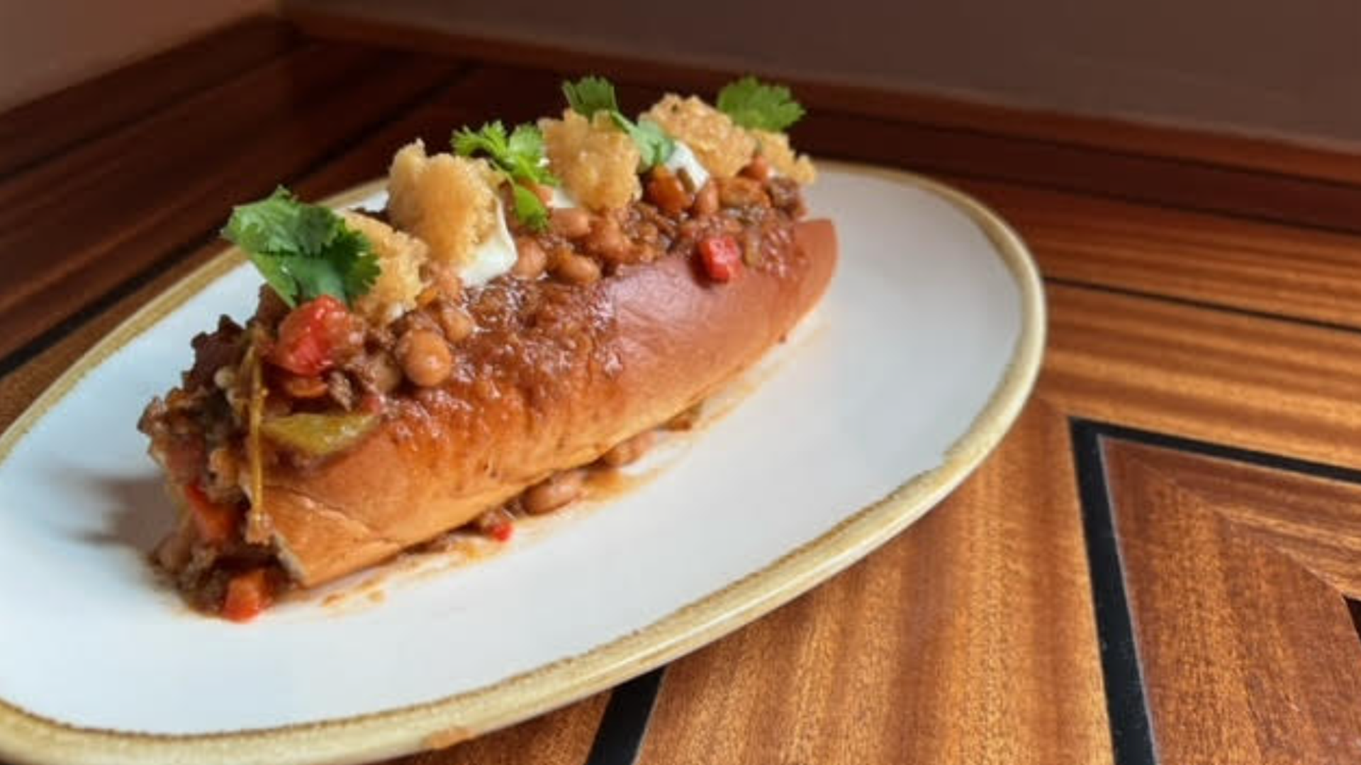 A hot dog loaded with mozzarella, bolognese sauce and fried garlic, topped with herbs is on a white plate.