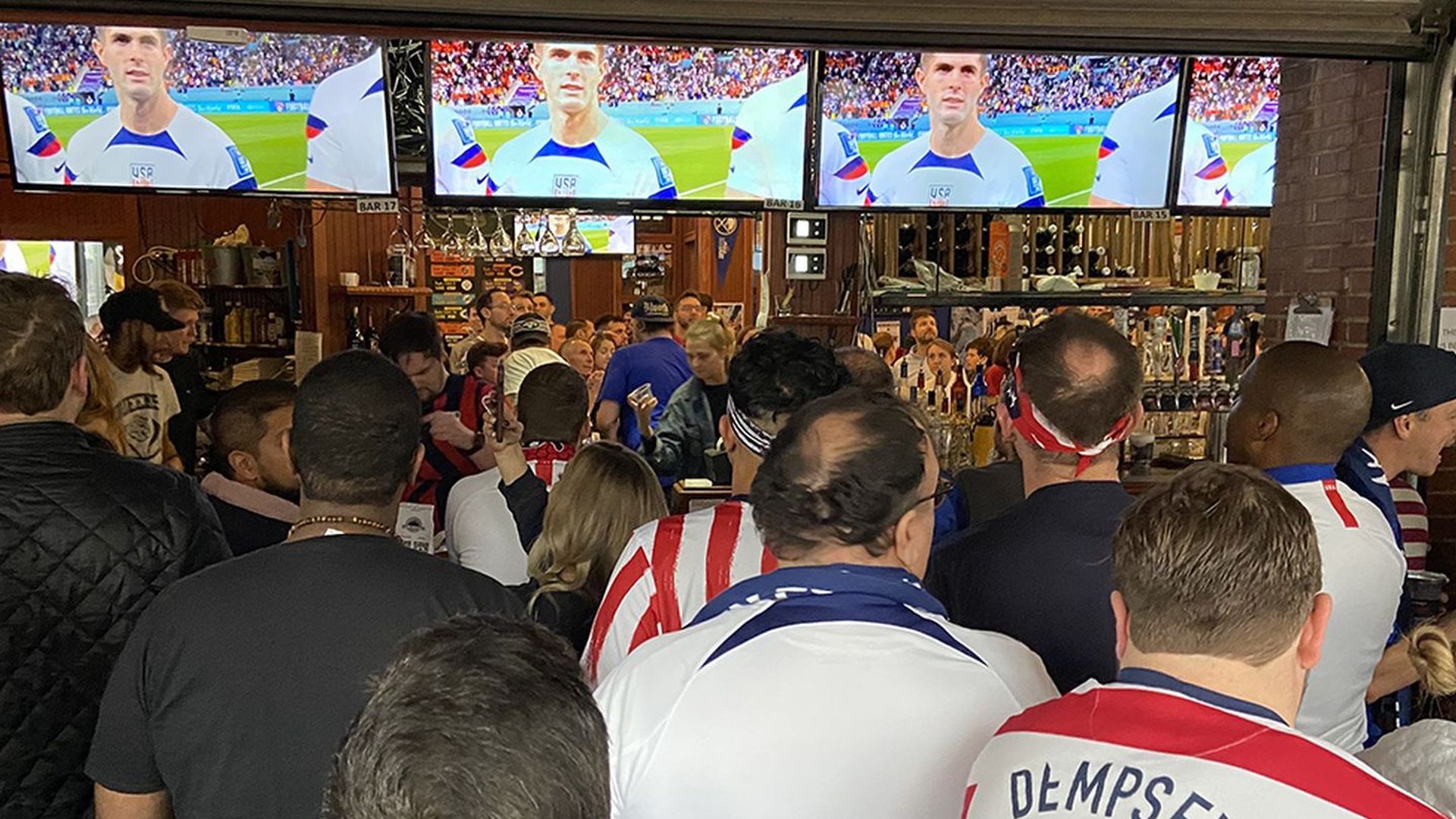 Busy sports bar with four large screens showing a soccer player in a white and blue USA jersey. Crowded patrons wear red, white, and blue; brick walls, bar taps, and busy shelves in the background.