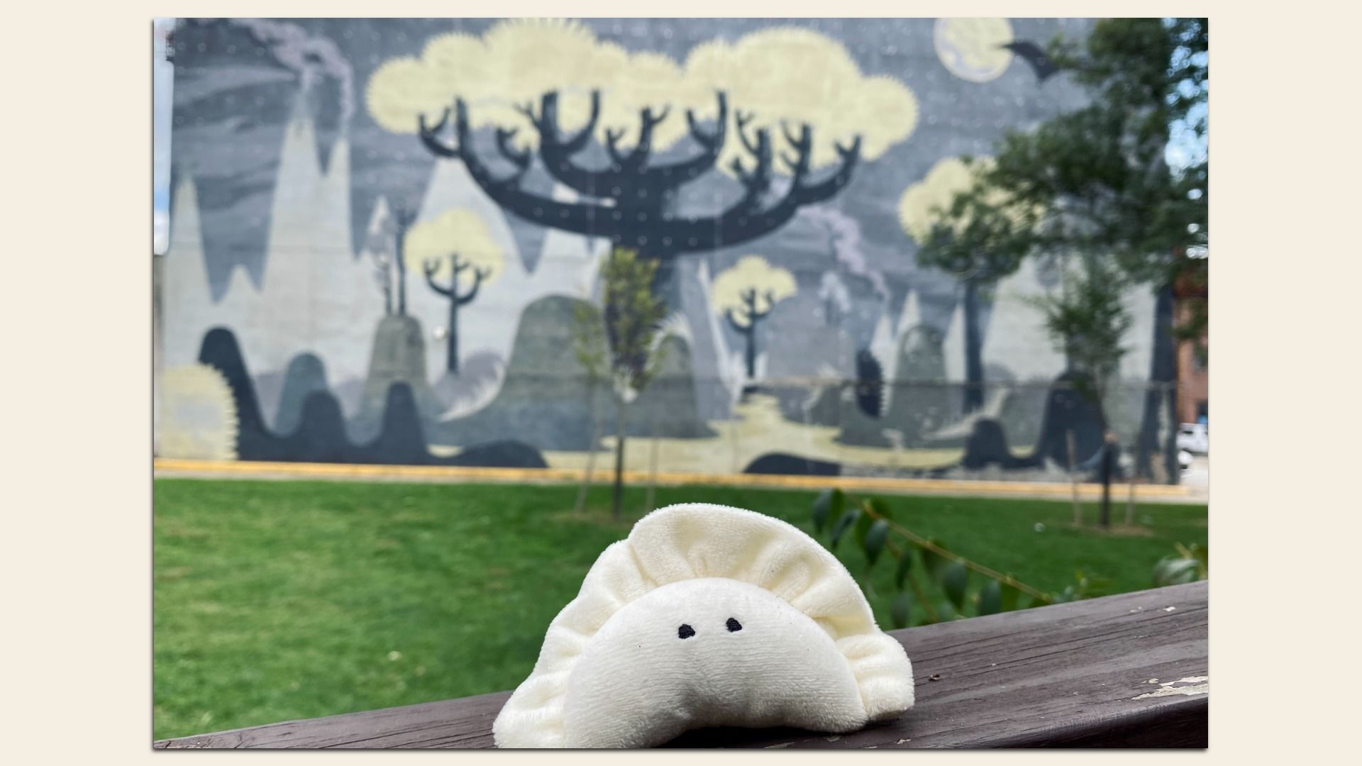 White plush dumpling with black eyes on a wooden railing, green grass and abstract black-and-yellow mural of trees and mountains on a wall in the background under a cloudy sky.