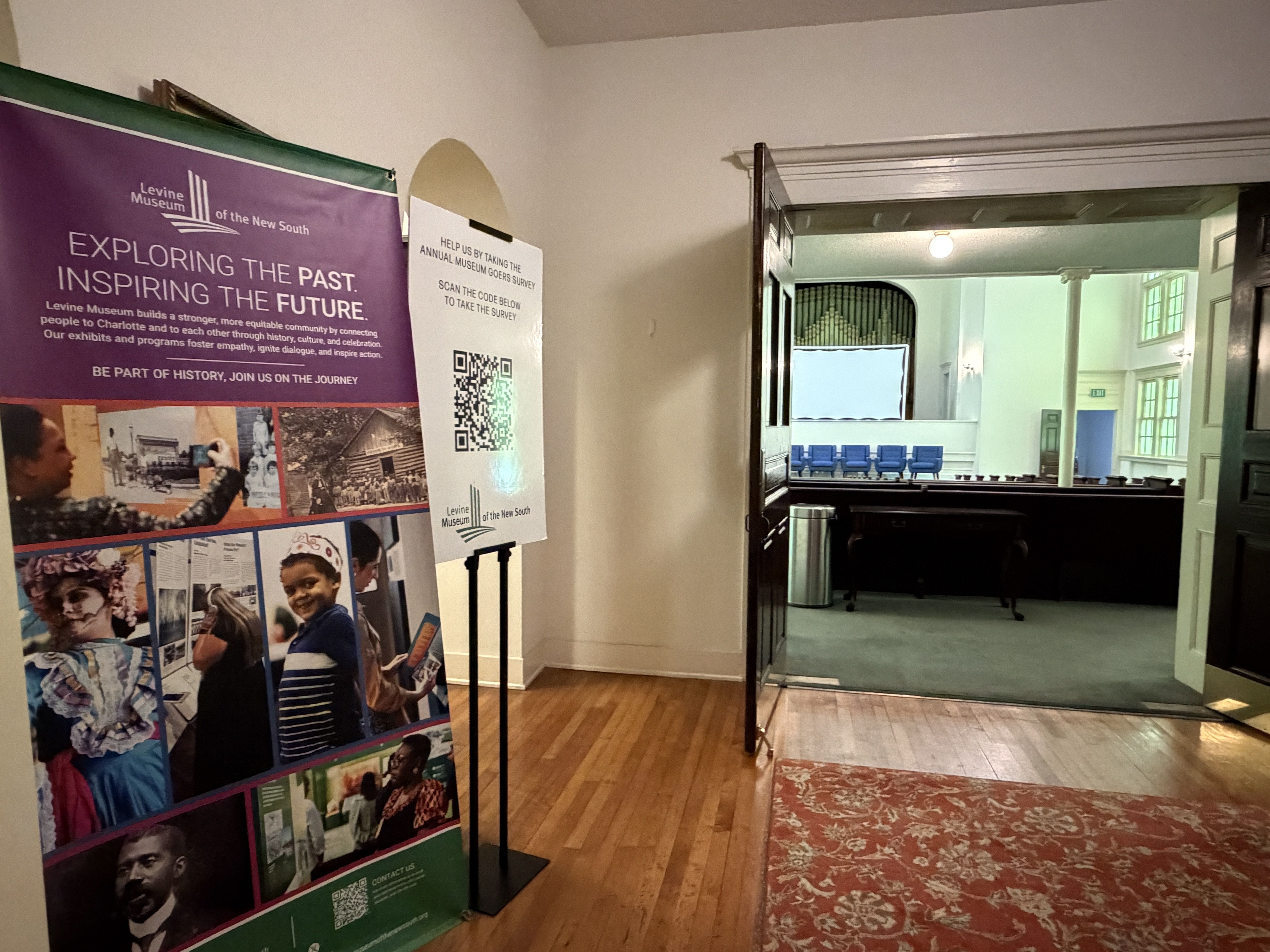 Museum hallway with a large purple banner reading "Levine Museum of the New South" and "EXPLORING THE PAST. INSPIRING THE FUTURE." A photo collage wall, wood floor, red rug, and a doorway to a room with blue chairs.