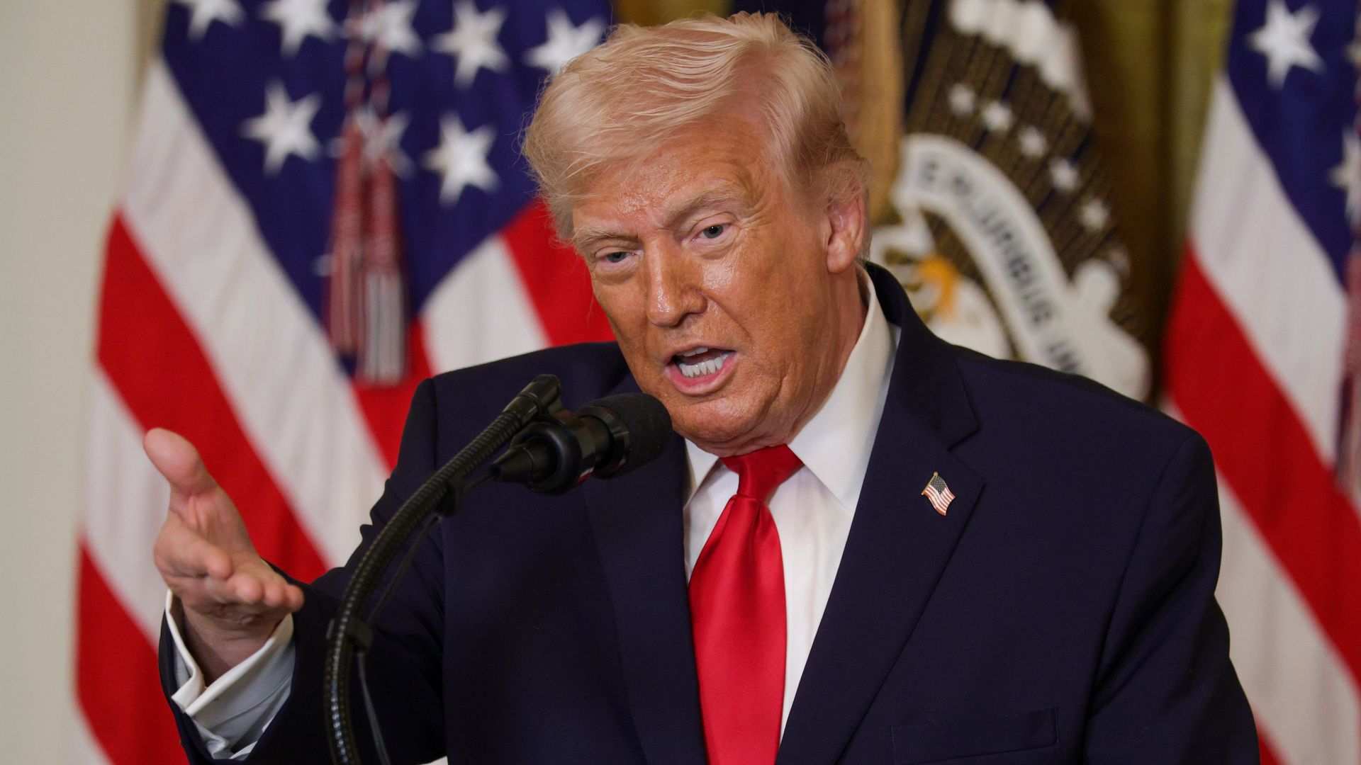 President Trump in a navy suit and red tie speaks into a microphone, gesturing with his hand, with American flags in the background.