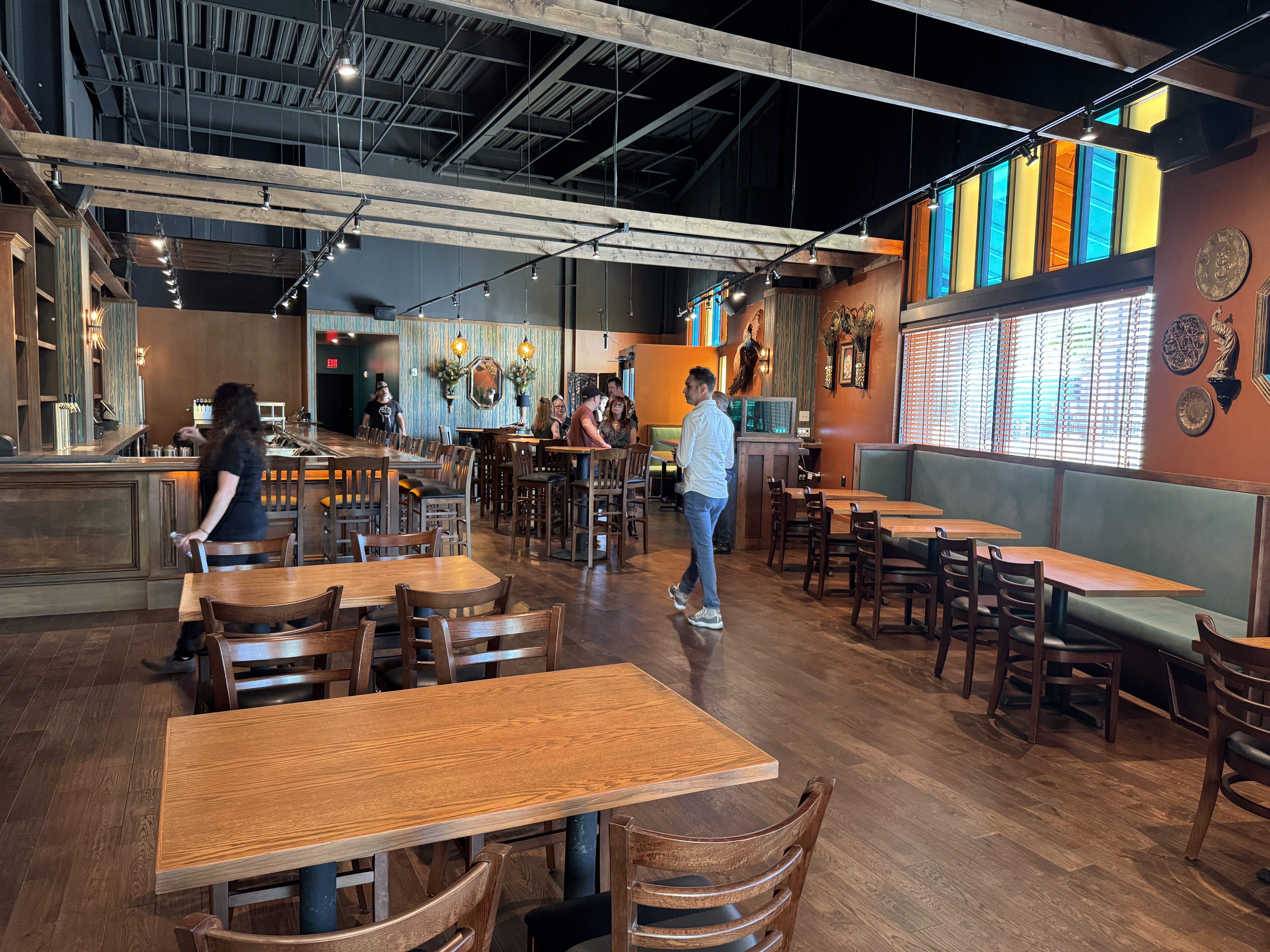 Spacious restaurant interior with wooden tables and chairs, stained glass windows in blue and orange, decorative wall plates, and people standing near the bar and tables.