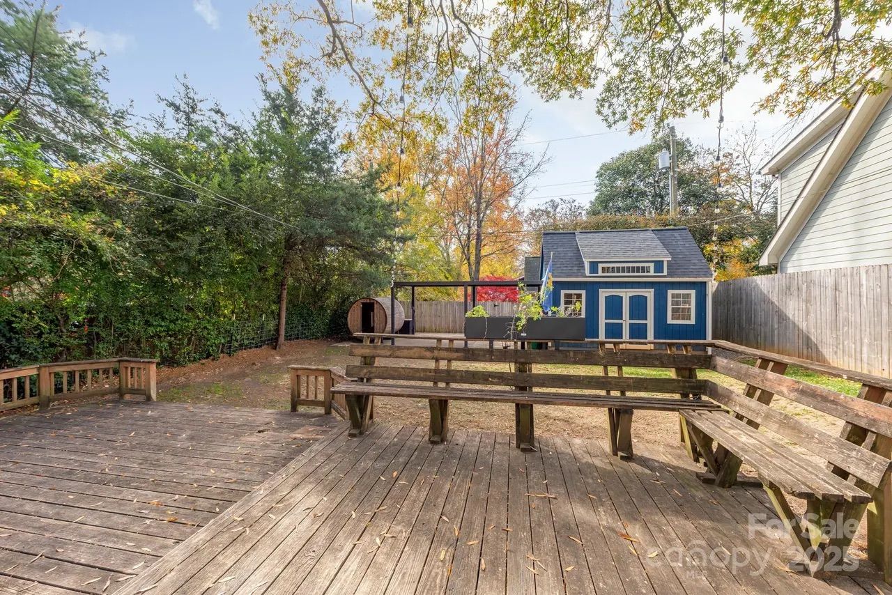 Backyard with large wooden deck featuring built-in benches, surrounded by trees with autumn leaves, a blue garden shed, and a round wooden structure near a wooden fence.