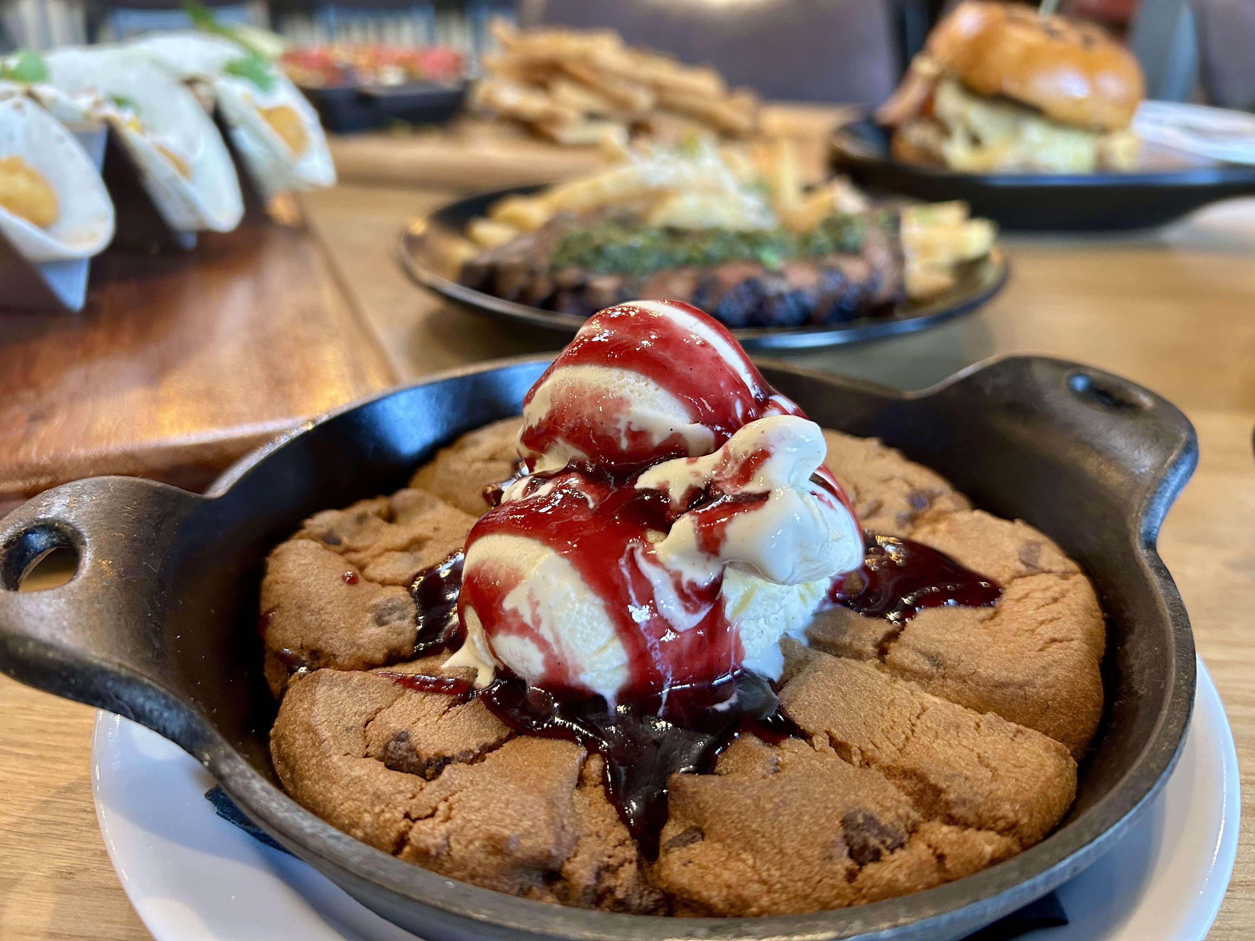heist brewery. Cast-iron skillet cookie topped with vanilla ice cream and red berry sauce; blurred background shows a burger with fries and wraps on a wooden table.