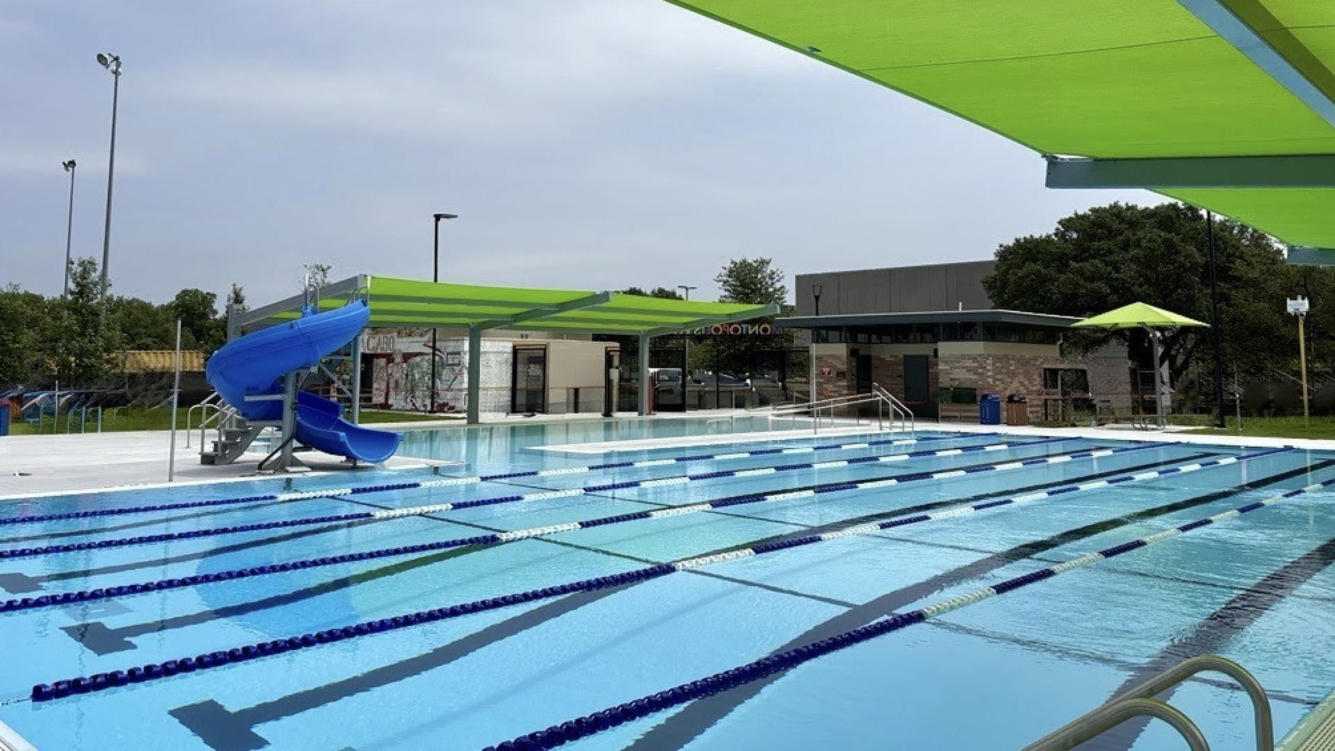 Outdoor swimming pool with a blue water slide on the left, bright lime-green shade sails overhead, blue-lane divided pool, and a building with trees in the background.