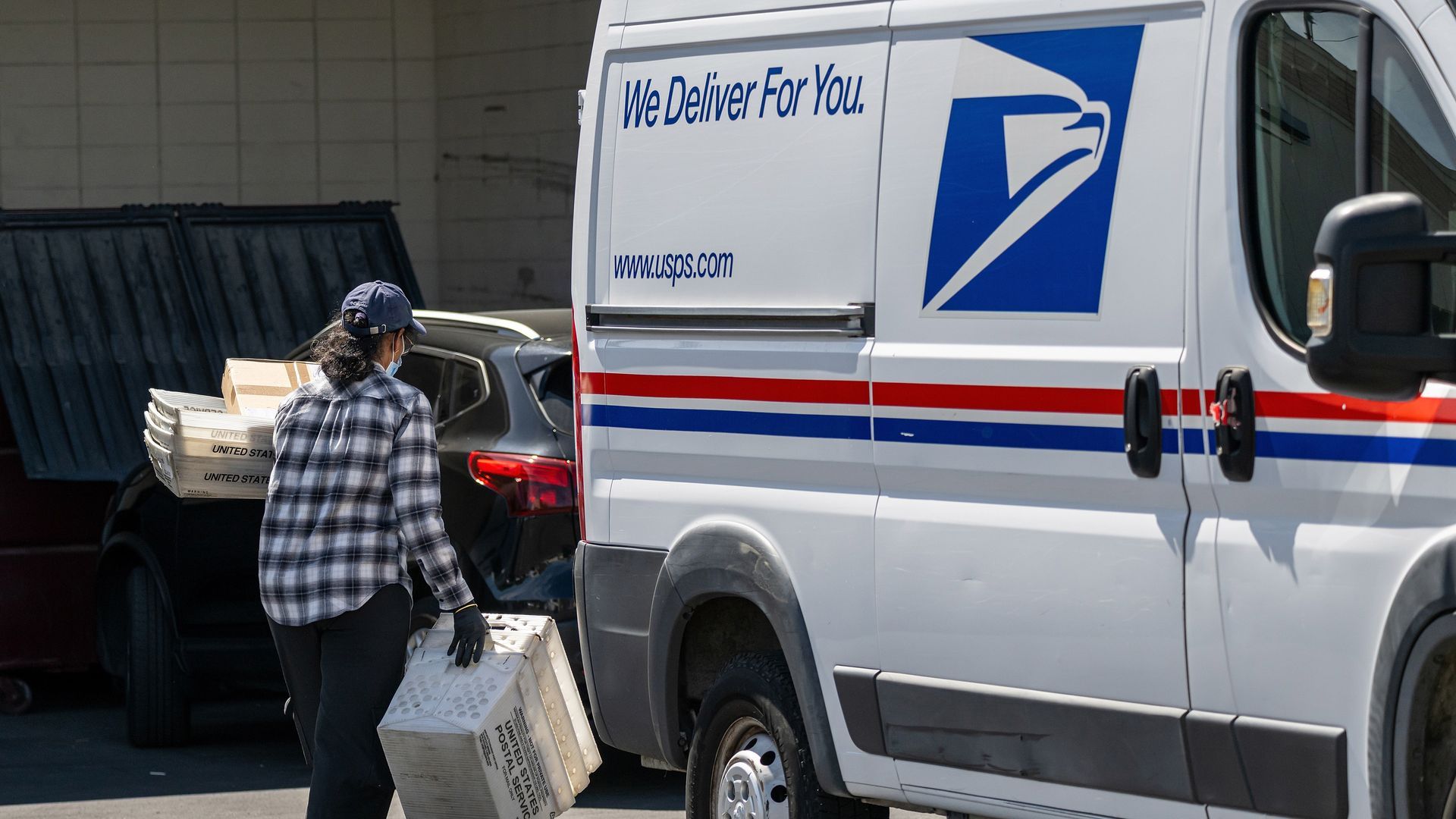 USPS delivery van with red and blue stripes and the slogan "We Deliver For You"; a worker in a plaid shirt and cap loads boxes from a car into the van.